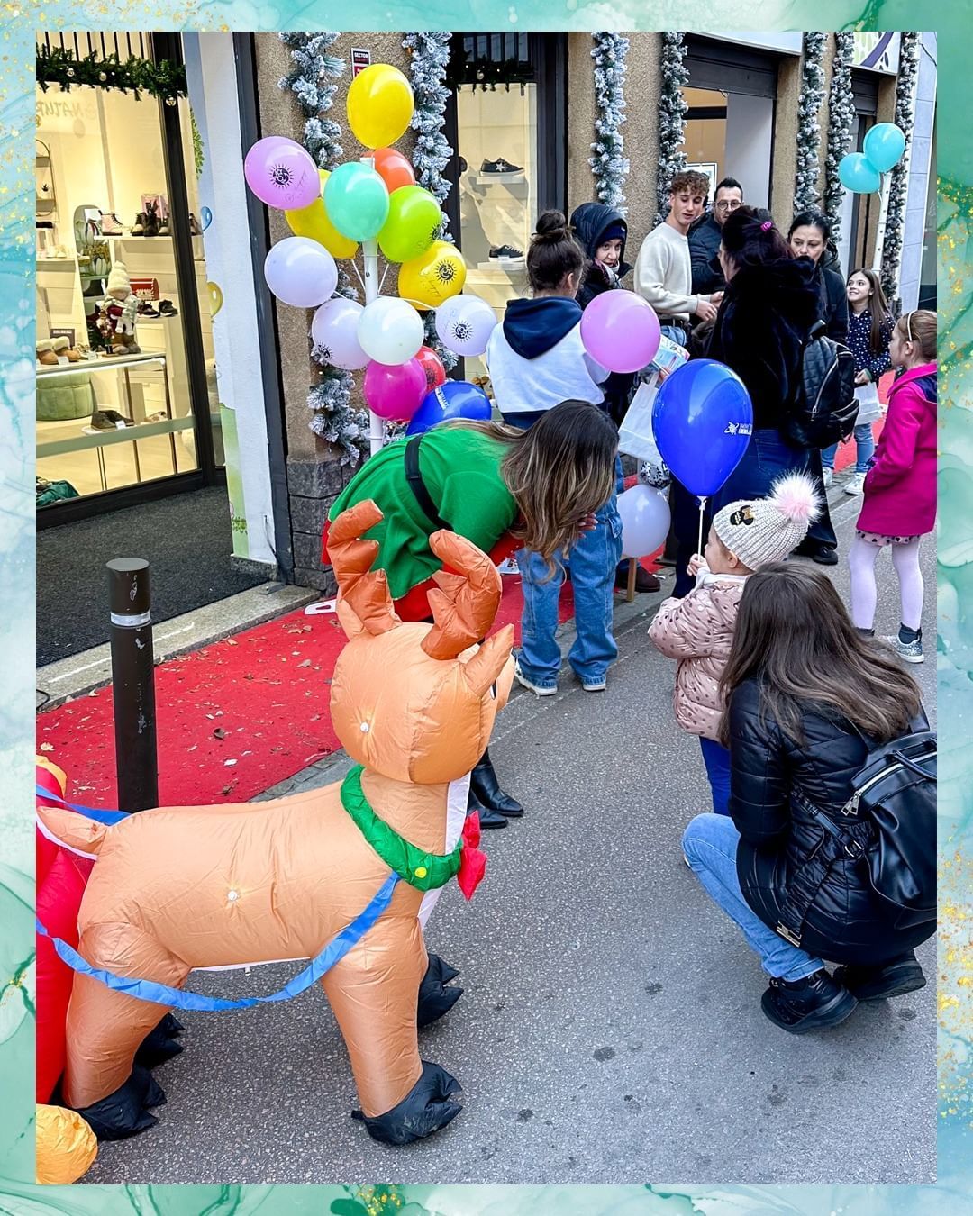 I bambini interagiscono con una renna gonfiabile e palloncini fuori da un negozio, con persone sullo sfondo.