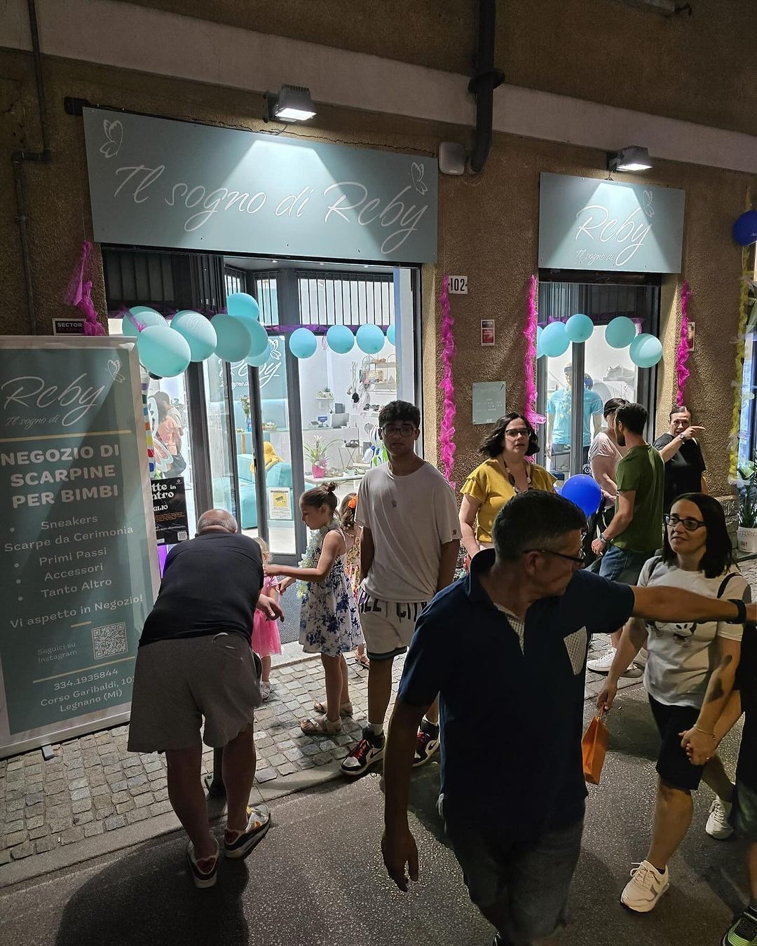 Persone fuori da un negozio decorato con palloncini blu e un cartello in italiano.