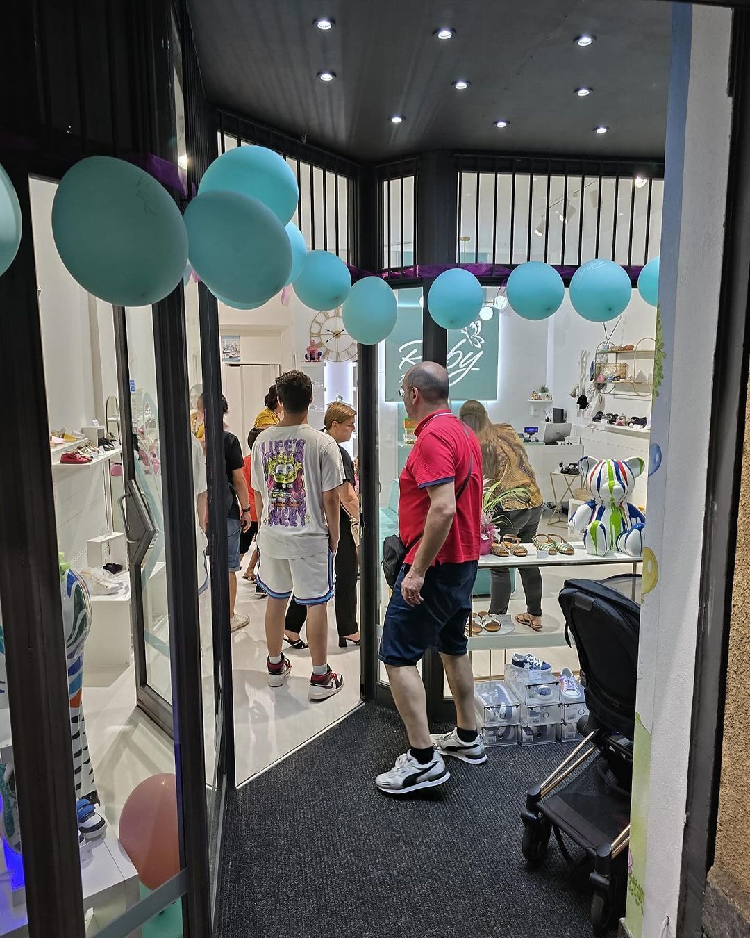 Persone all'interno di un negozio, viste dall'ingresso. Palloncini turchesi pendono dal soffitto. Entra un uomo con una camicia rossa.