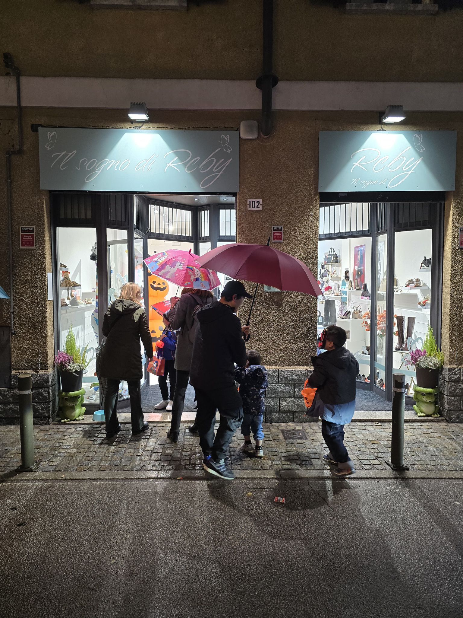 Persone con ombrelli entrano in un negozio in una notte piovosa. La vetrina ha grandi vetrate e illuminazione dall'alto.