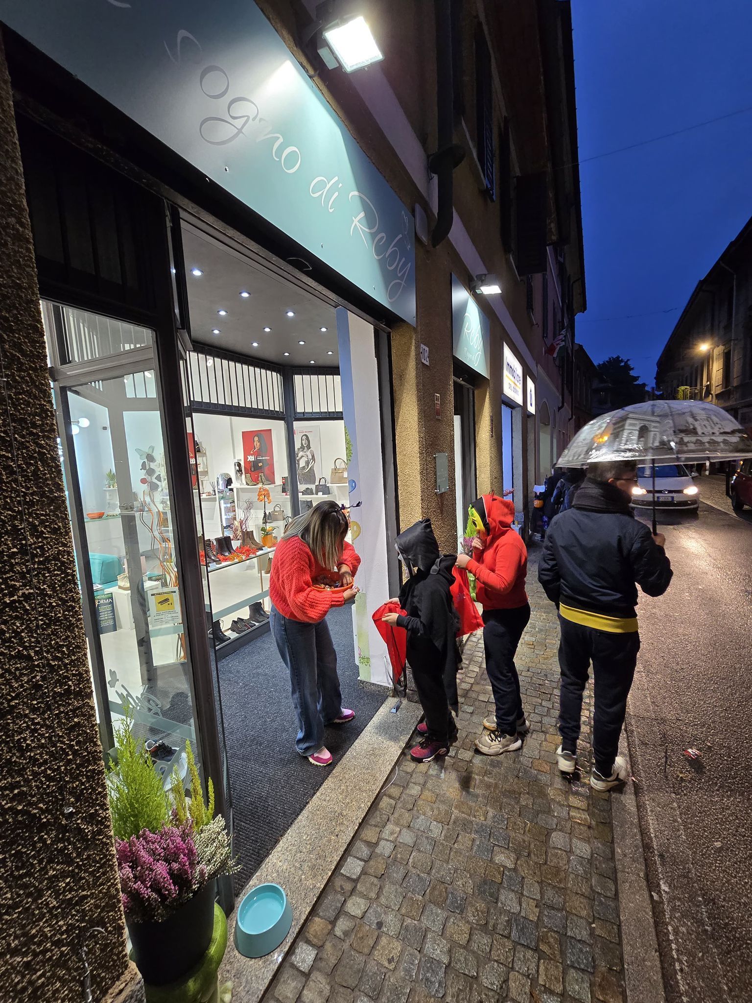 Persone fuori dalla vetrina di un negozio al tramonto. Alcune guardano dentro, indossando abiti rossi e neri.