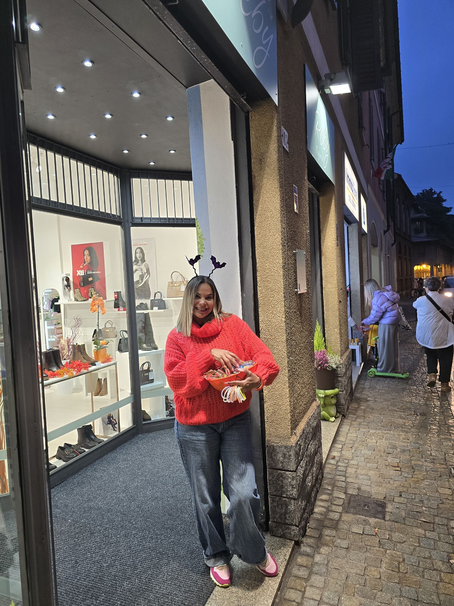Donna con maglione rosso fuori da un negozio, con in mano una ciotola di caramelle. Indossa jeans, scarpe rosa e un copricapo a forma di pipistrello.