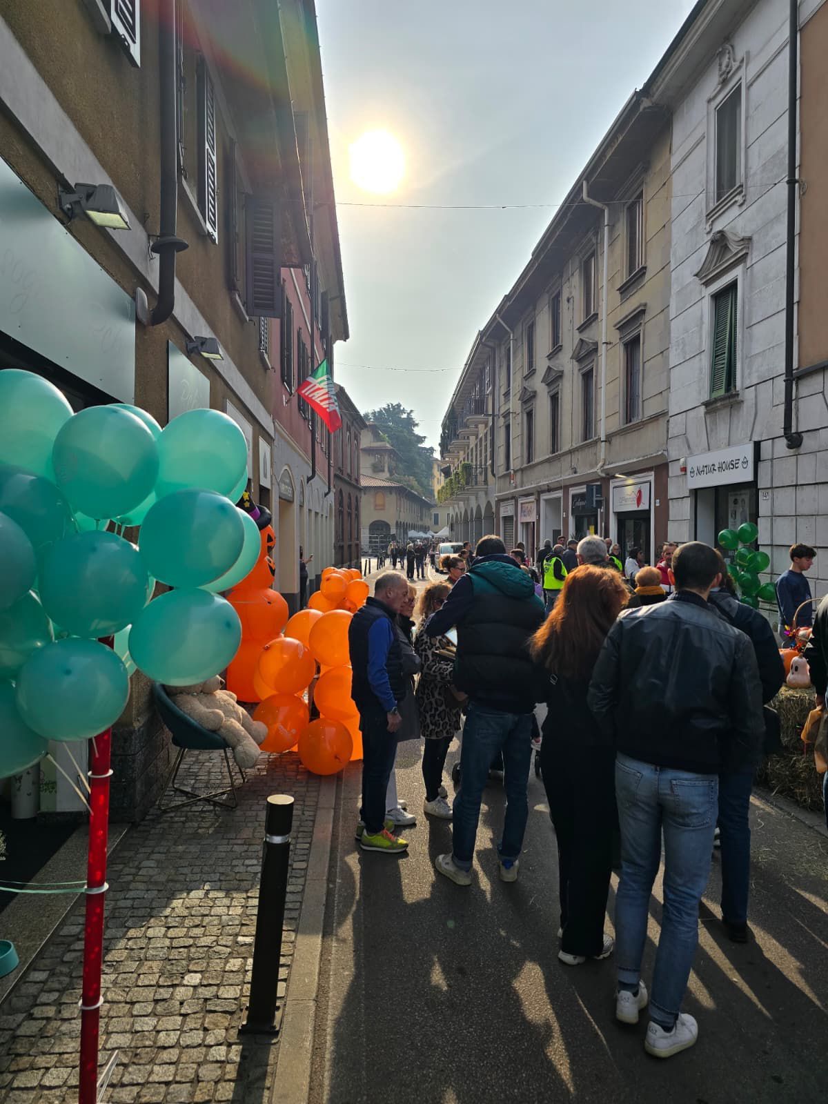 Scena di strada con pedoni e palloncini. Palloncini arancioni e verdi, edifici e un cielo soleggiato.