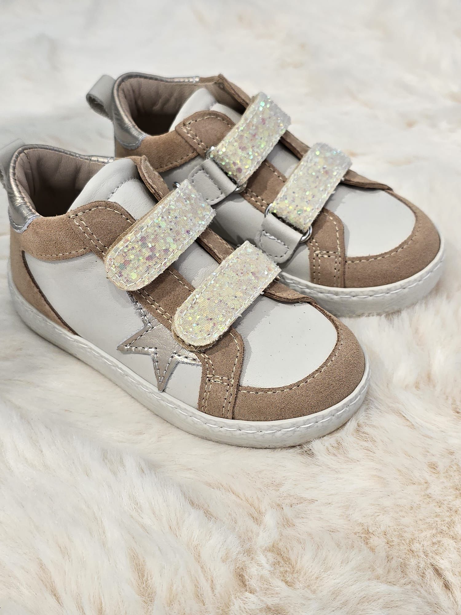 Sneakers per bambini color beige e bianco con cinturini glitterati e un dettaglio a forma di stella su uno sfondo soffice e chiaro.