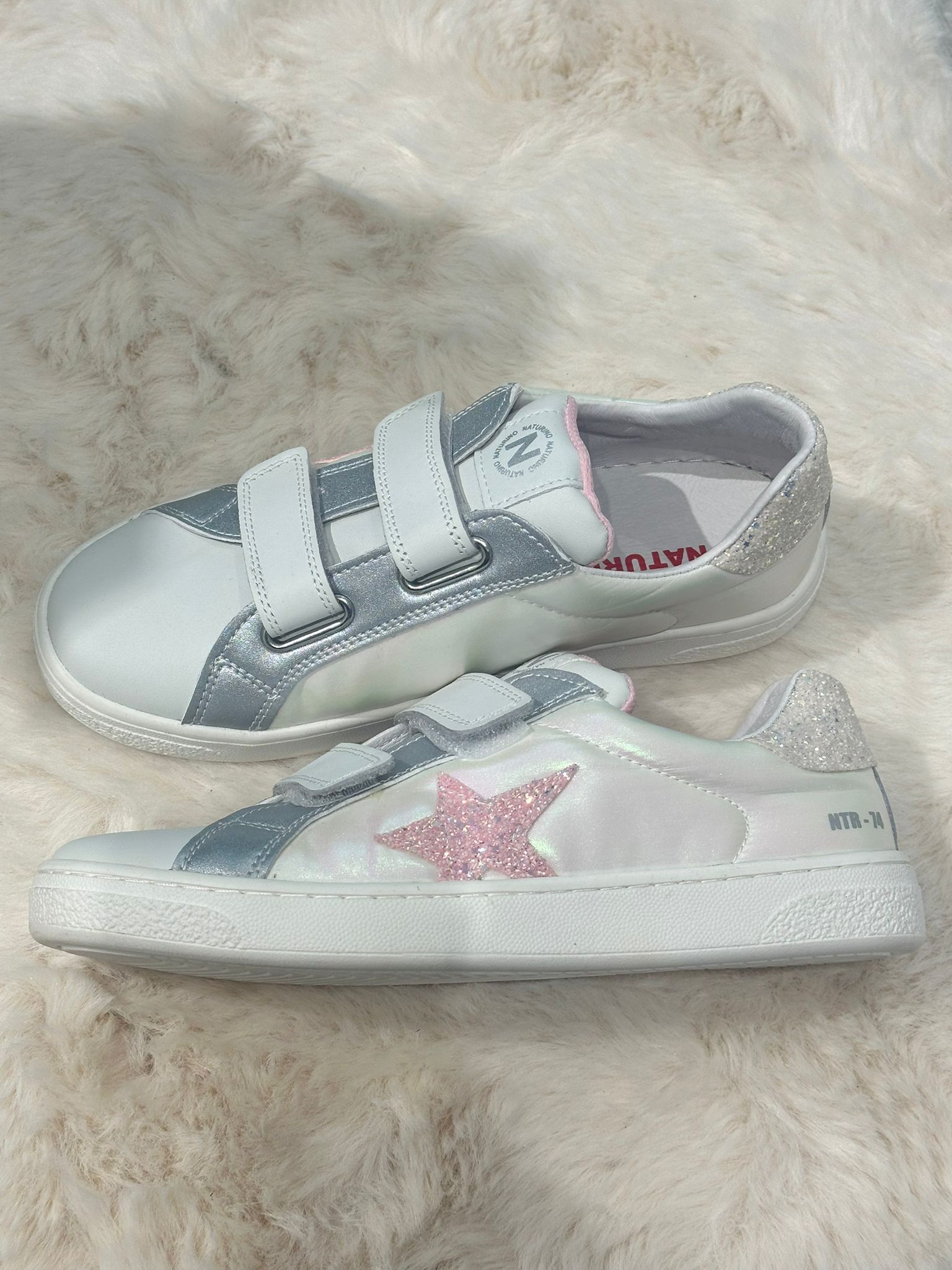 Sneakers bianche e argentate con cinturini in velcro e stella glitterata rosa su superficie sfocata.