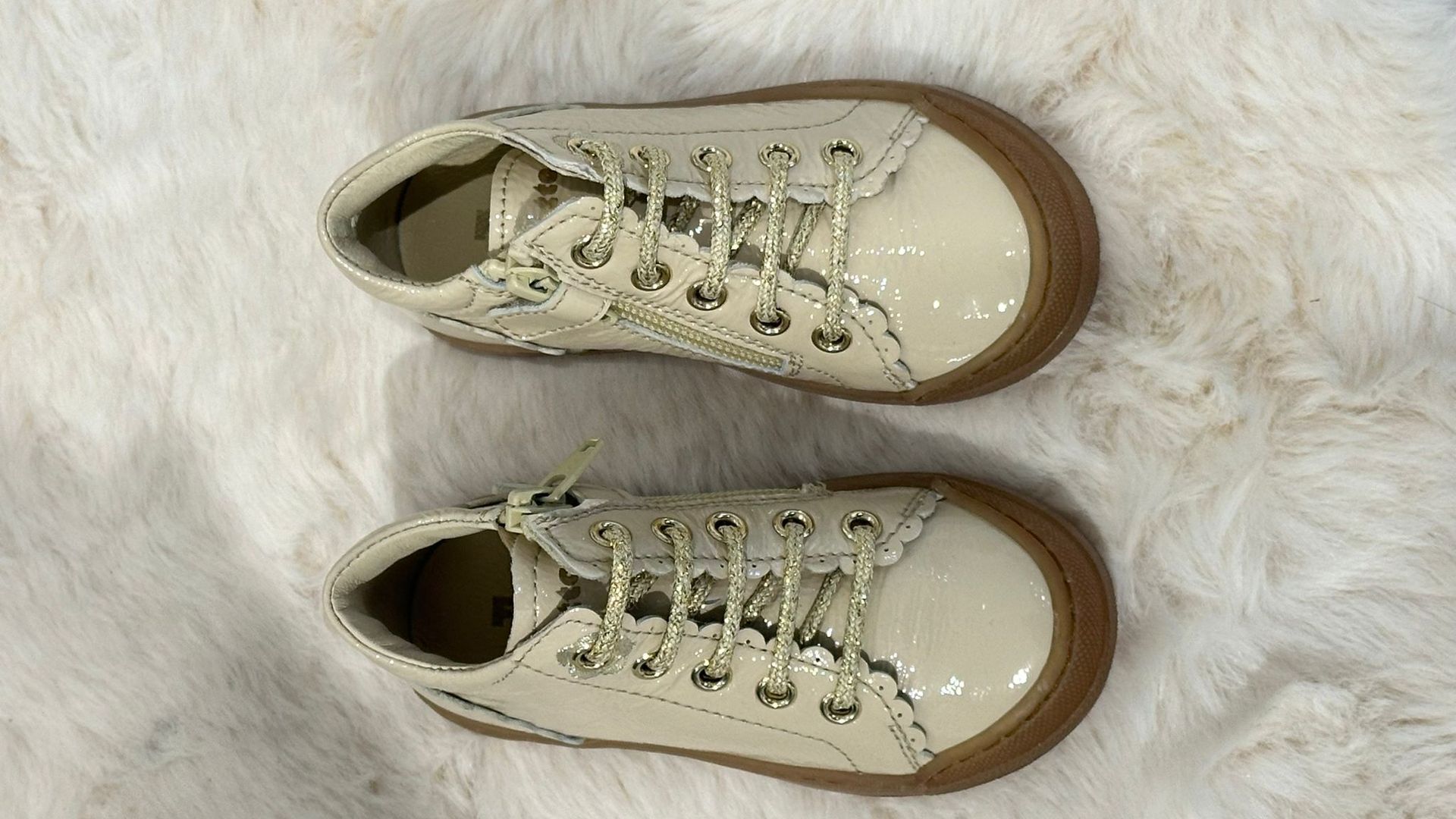 Scarpe da ginnastica color crema per bambini, con suole e lacci marroni, appoggiate su una soffice superficie bianca.