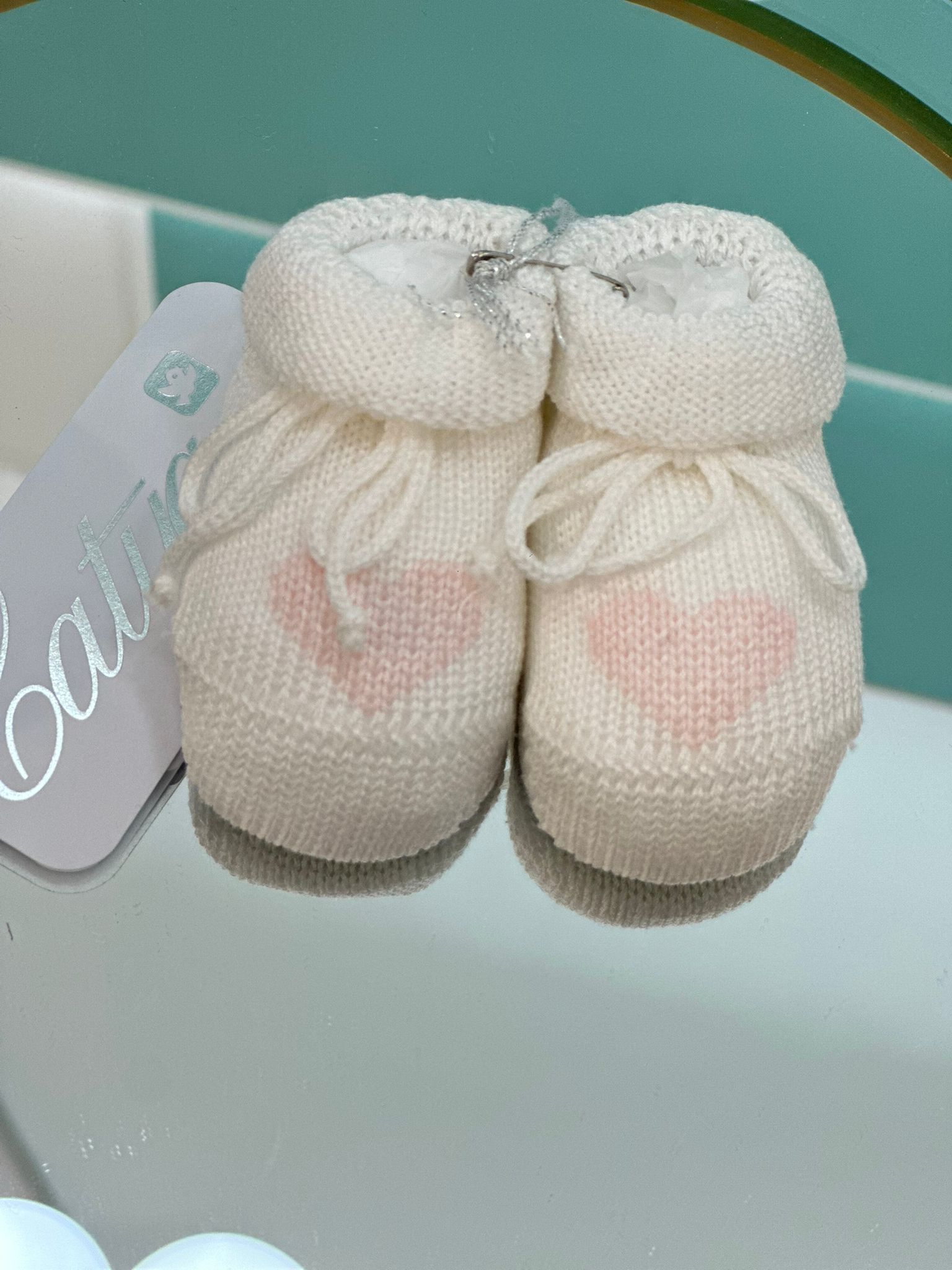Scarpine bianche per neonati lavorate a maglia con cuori rosa, su superficie riflettente.