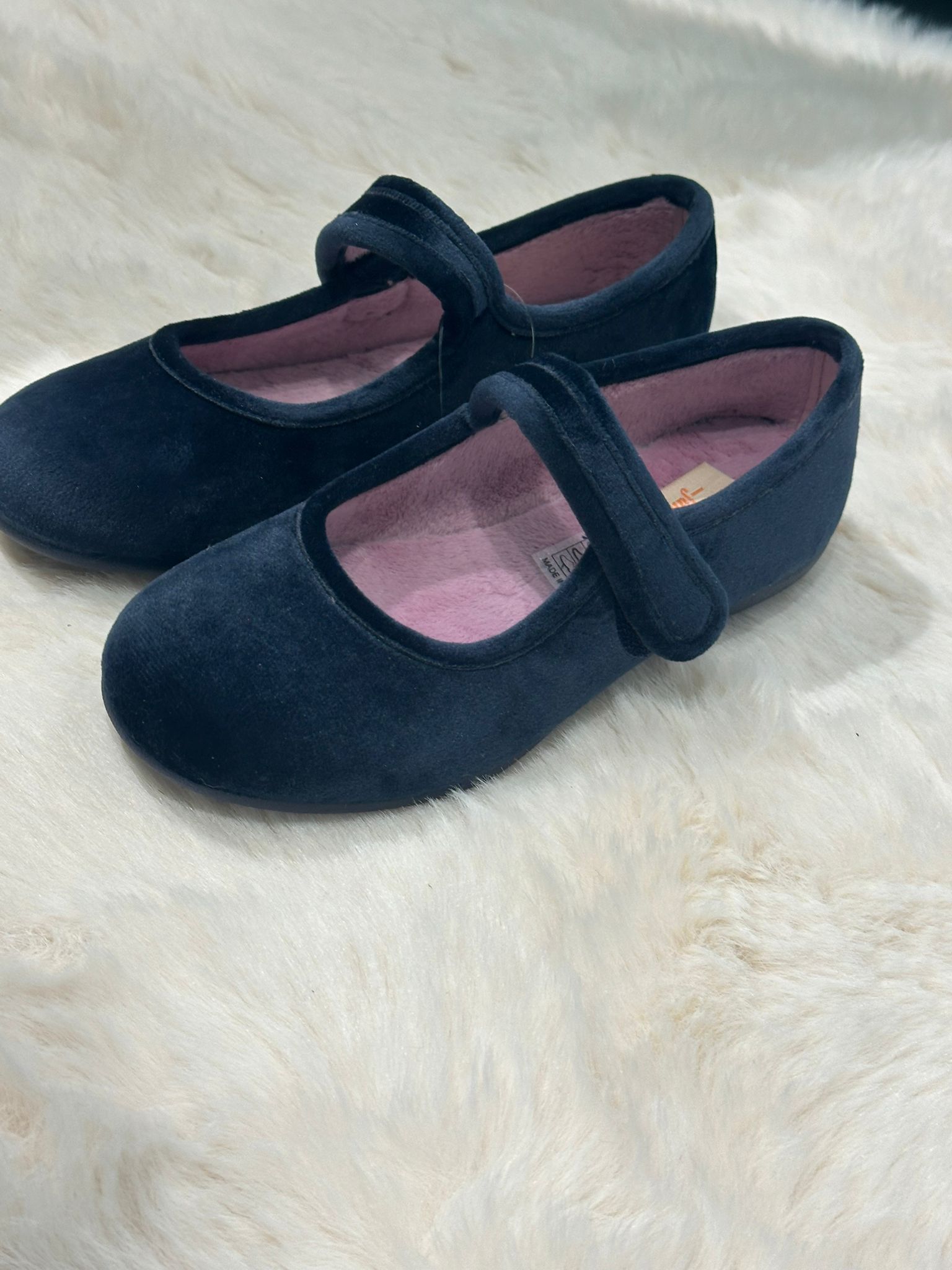 Un paio di scarpe blu scuro in stile Mary Jane con cinturini in velcro, su una superficie bianca e pelosa.