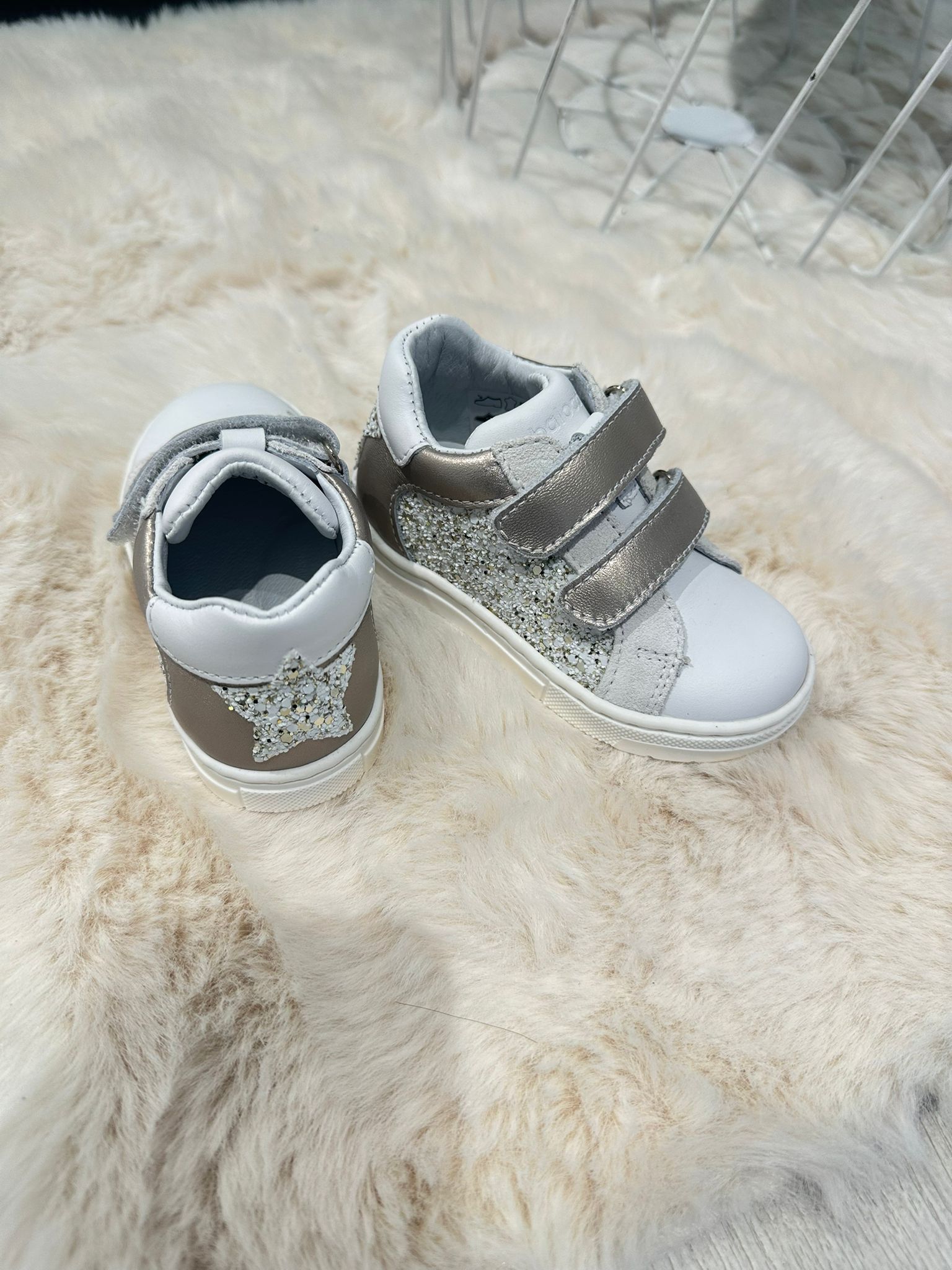 Sneakers per bambini bianche e argento con dettagli a stella e cinturini a strappo, su un soffice tappeto.
