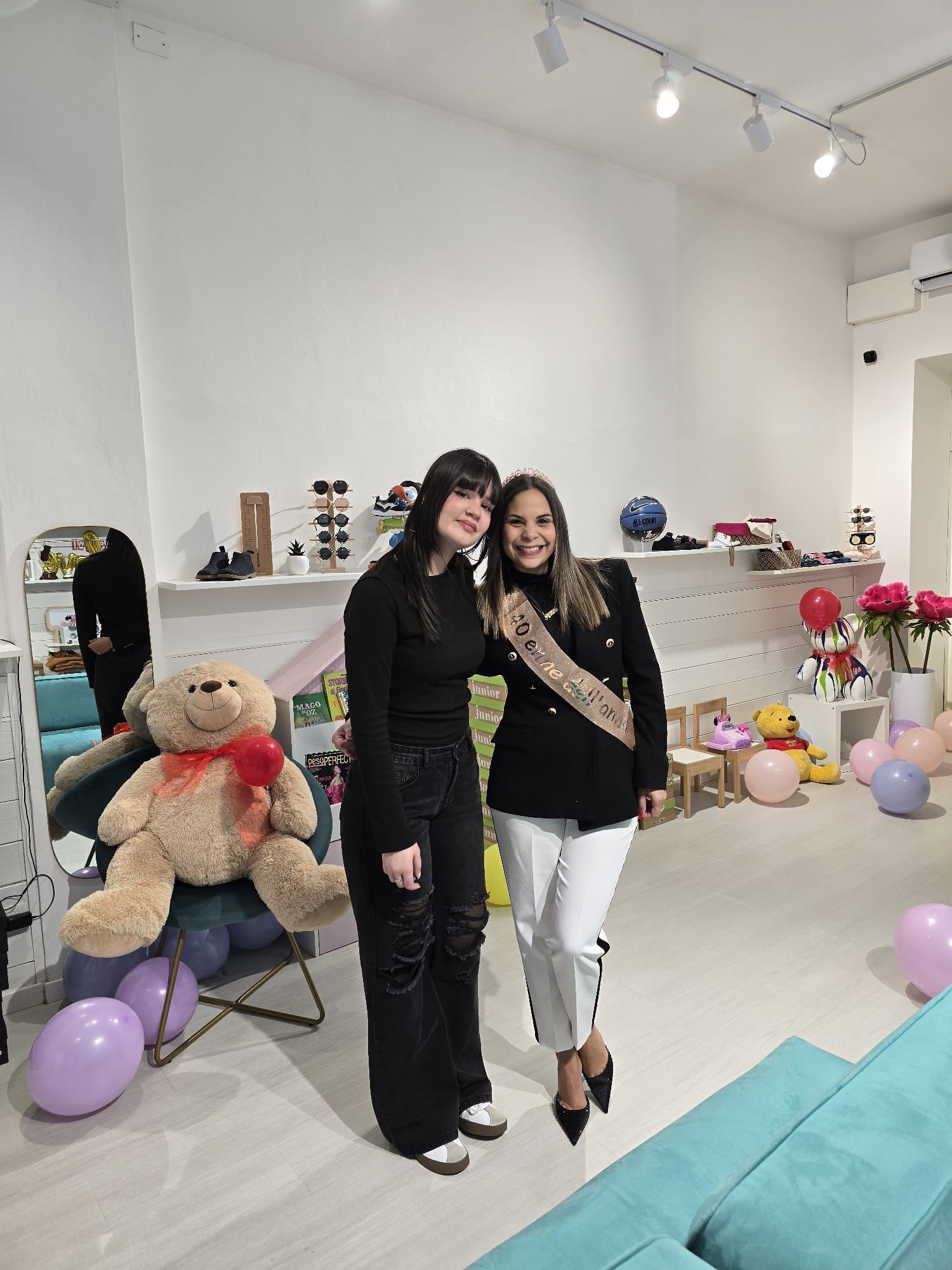 Due donne posano in una boutique illuminata, una delle quali indossa una fascia, circondate da articoli per bambini, palloncini e un orsacchiotto di peluche.