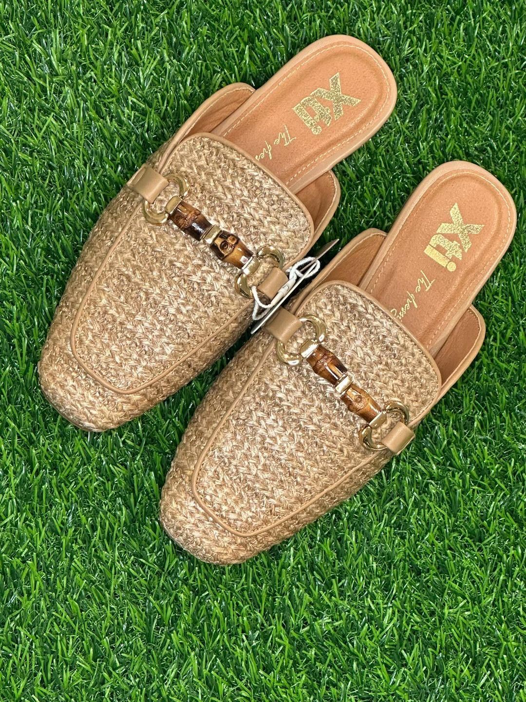 Un paio di mules slip-on intrecciate color cuoio con dettagli in stile bambù, su erba verde.