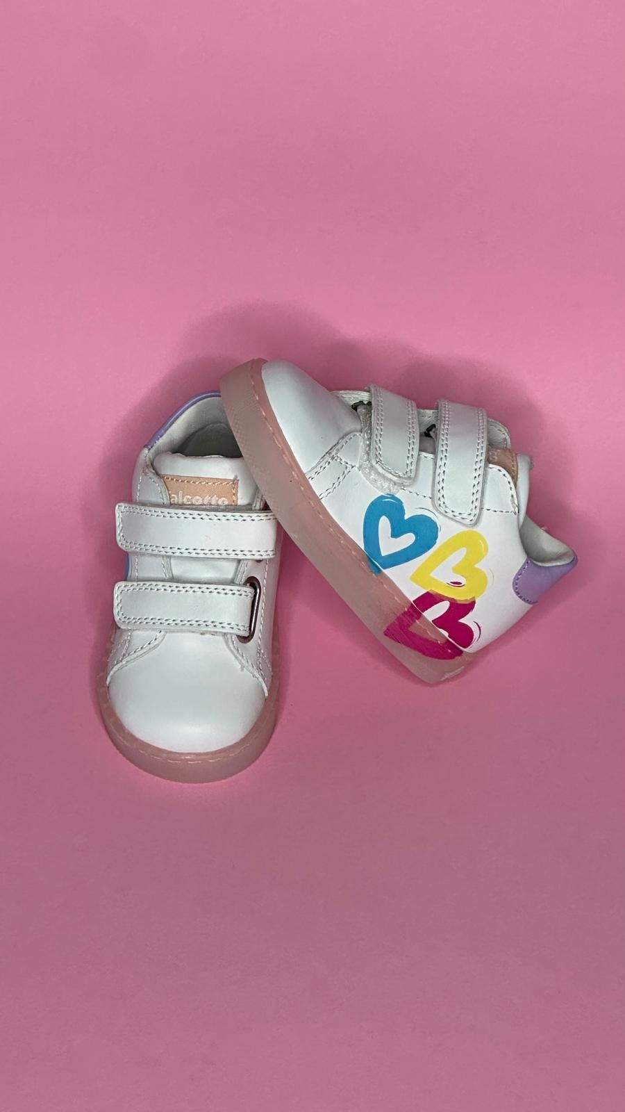 Scarpine bianche per bambini con disegno a cuore su sfondo rosa.