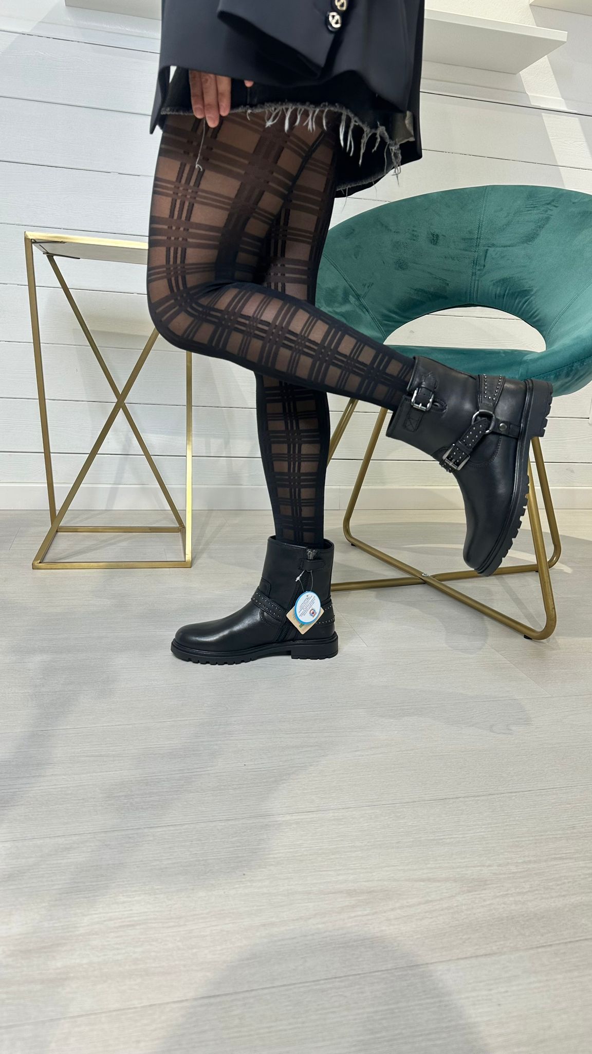 Persona che indossa collant fantasia e stivali neri, in posa vicino a un tavolo dorato e una sedia verde acqua.