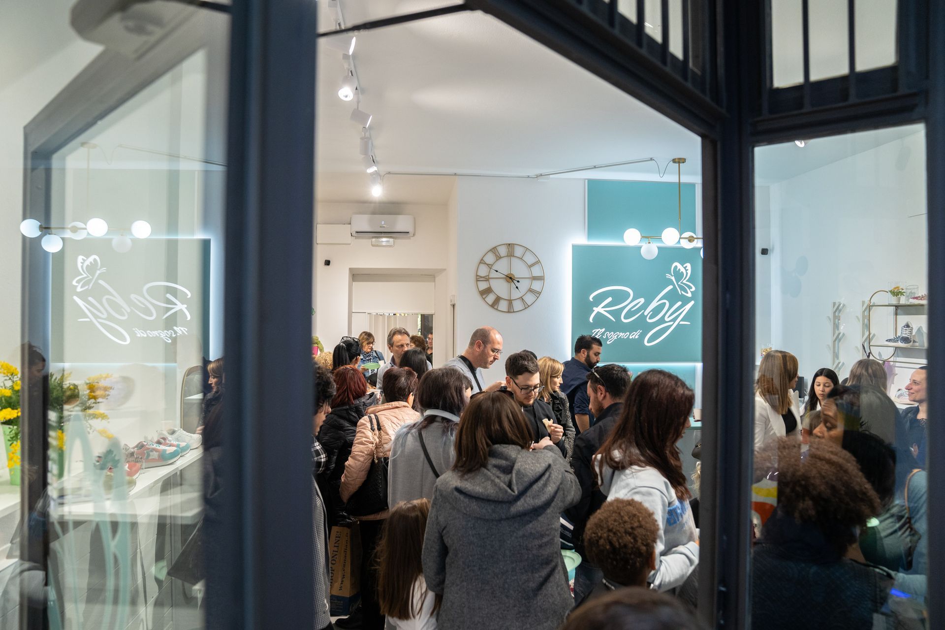 La gente si è radunata all'interno di un negozio con una parete verde acqua e un grande orologio. Attraverso una porta a vetri.