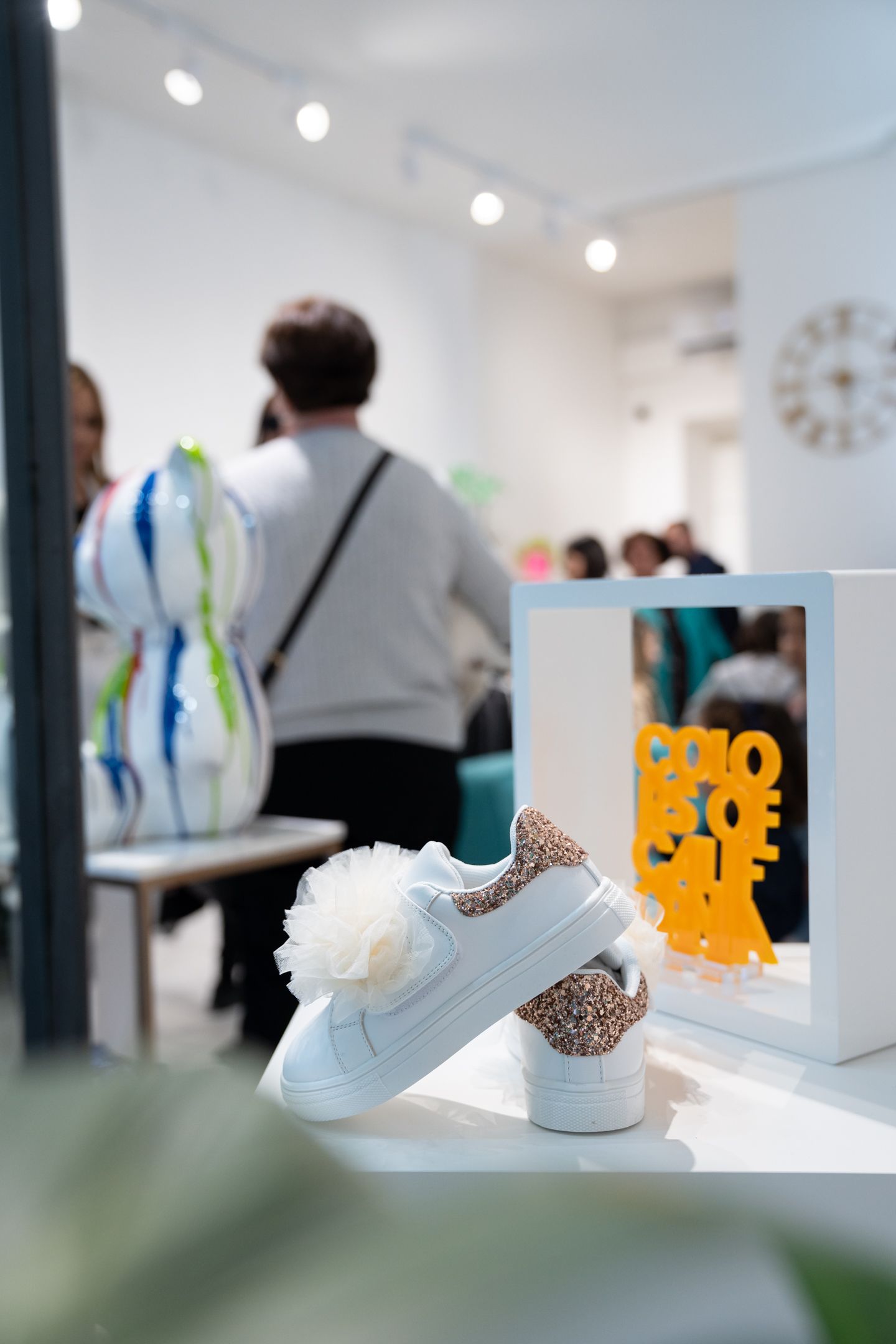 Sneakers bianche con pompon e stampa leopardata, esposte in una galleria con persone sullo sfondo.