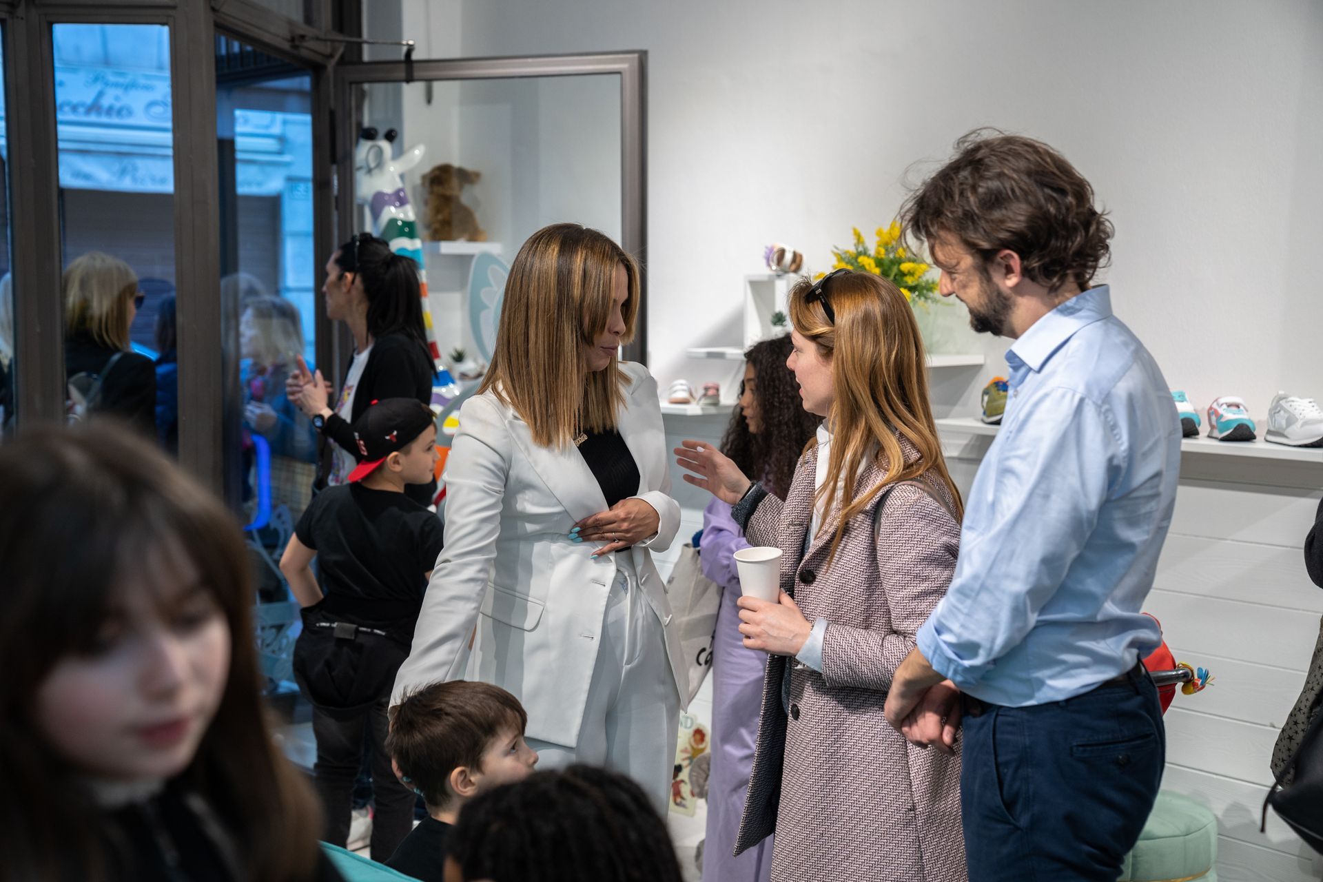 Persone che conversano in una galleria d'arte. Una donna in abito bianco parla con i visitatori, mentre altri ammirano le mostre.