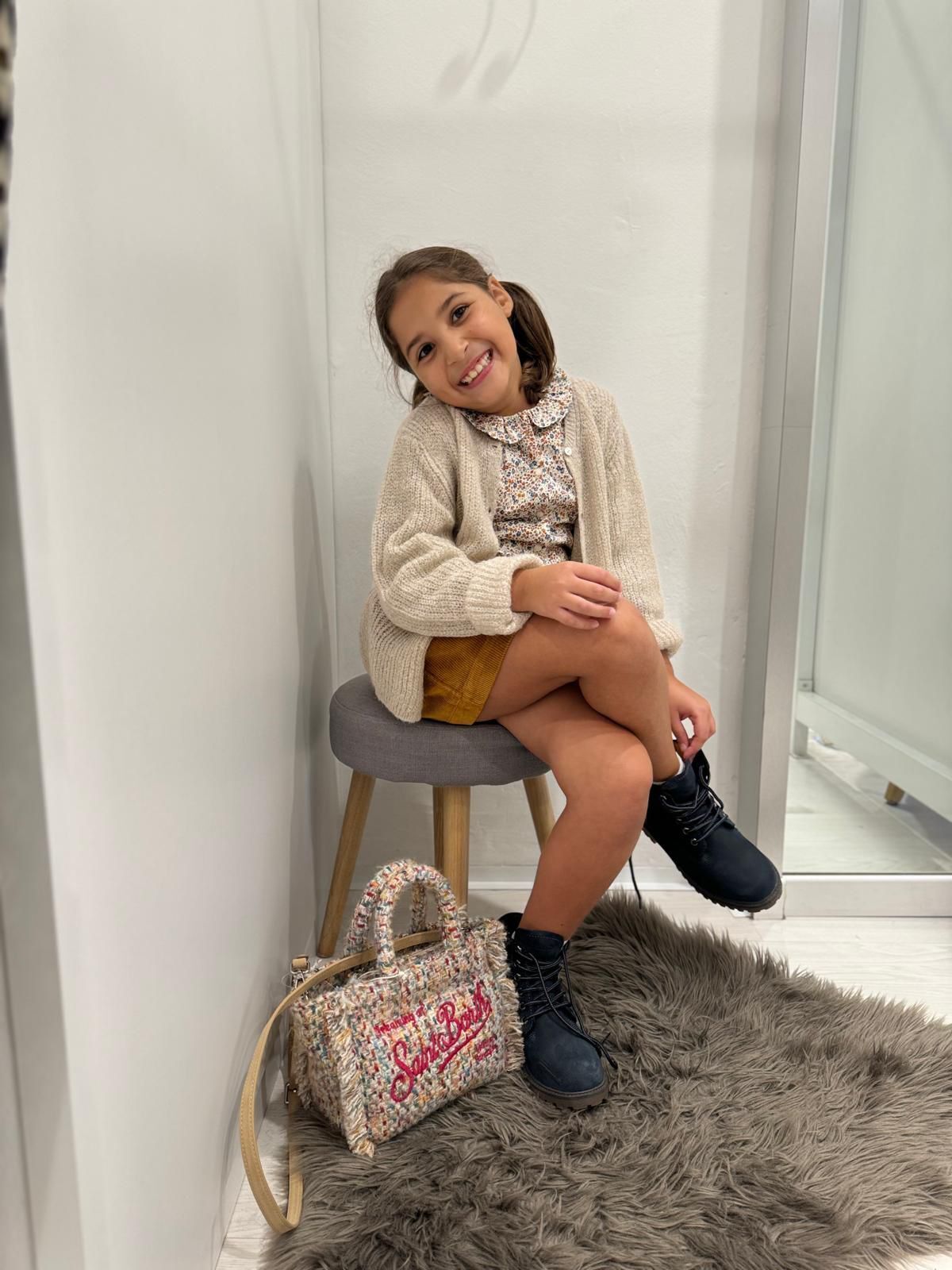 La ragazza sorride, seduta su uno sgabello nel camerino, indossa cardigan, gonna, stivali e tiene in mano una borsa.