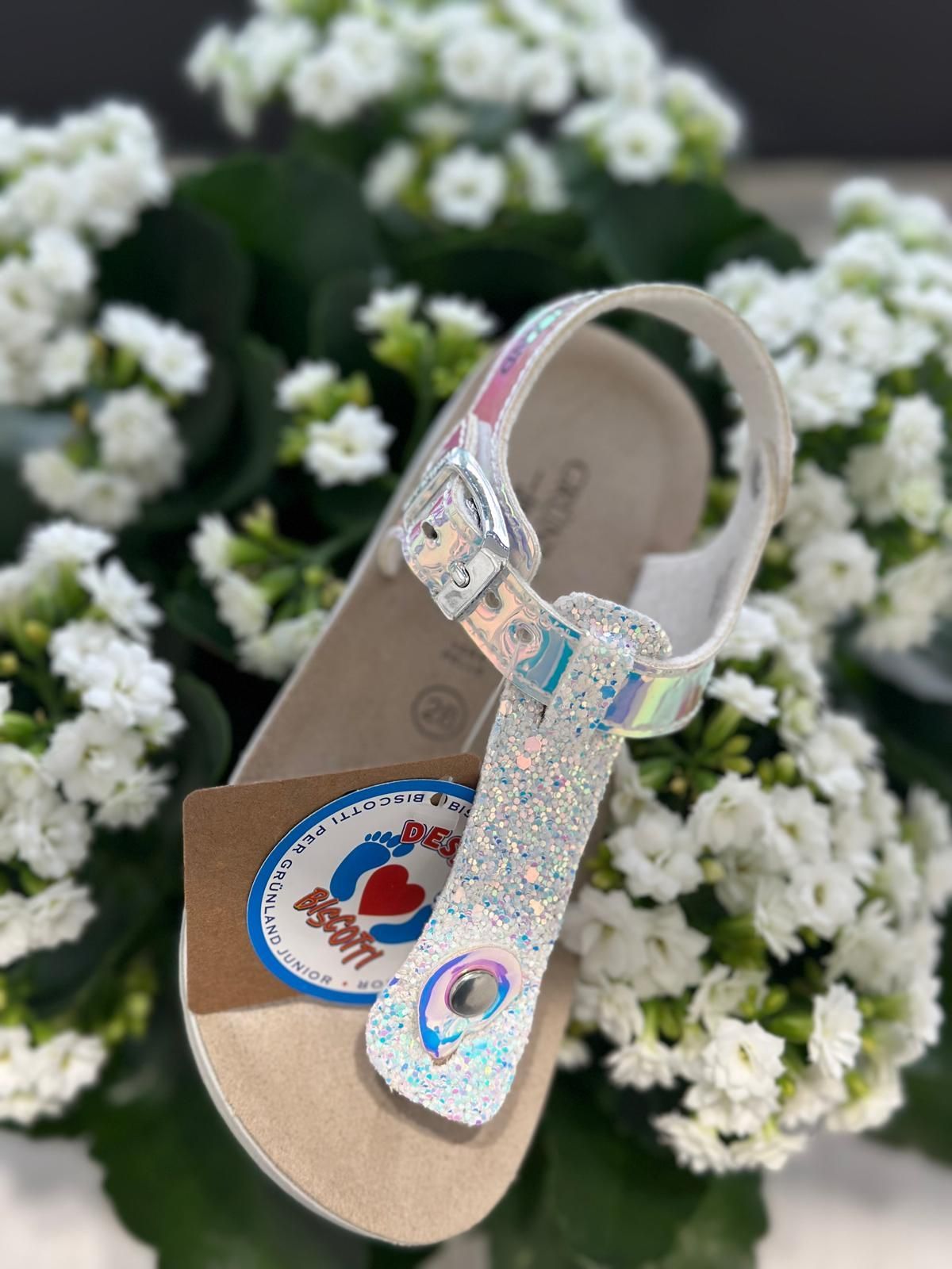 Sandalo bianco scintillante con dettagli olografici, appoggiato su fiori bianchi.