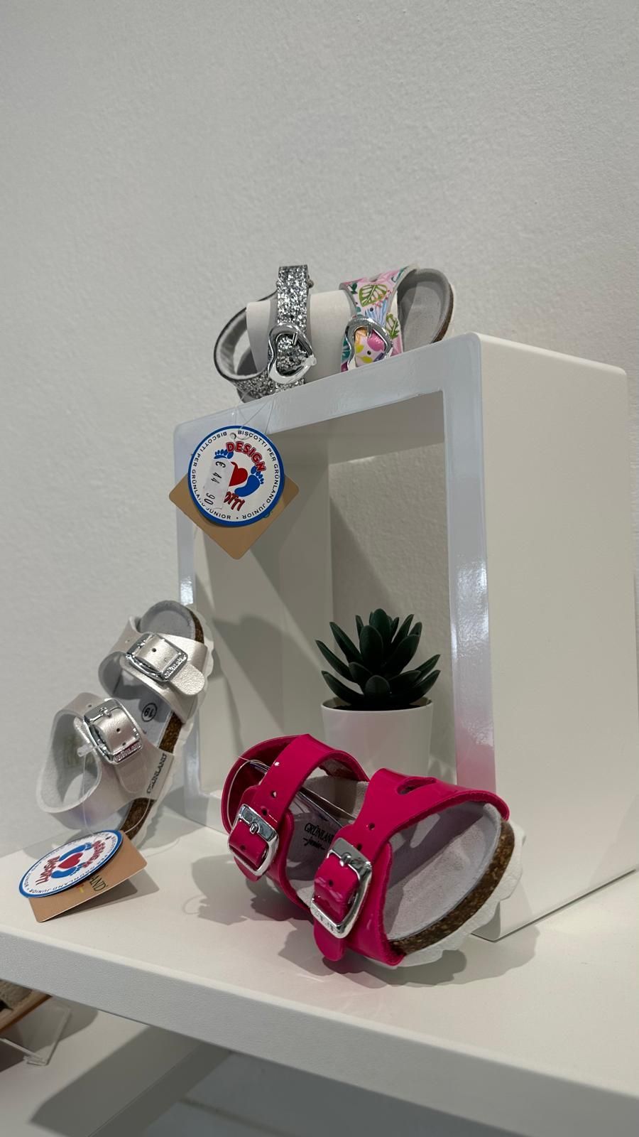 Sandali esposti su uno scaffale bianco. Tre paia, rosa, argento e floreale, con etichetta. Una piccola pianta sullo sfondo.