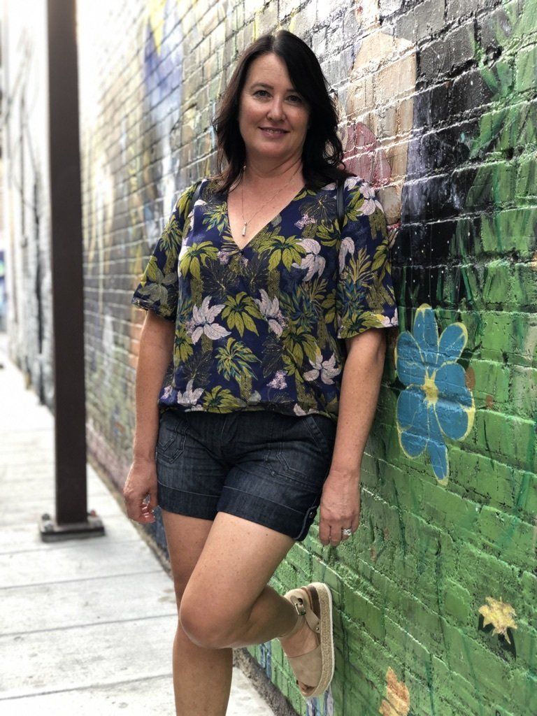 A woman in a floral shirt and shorts is leaning against a brick wall.