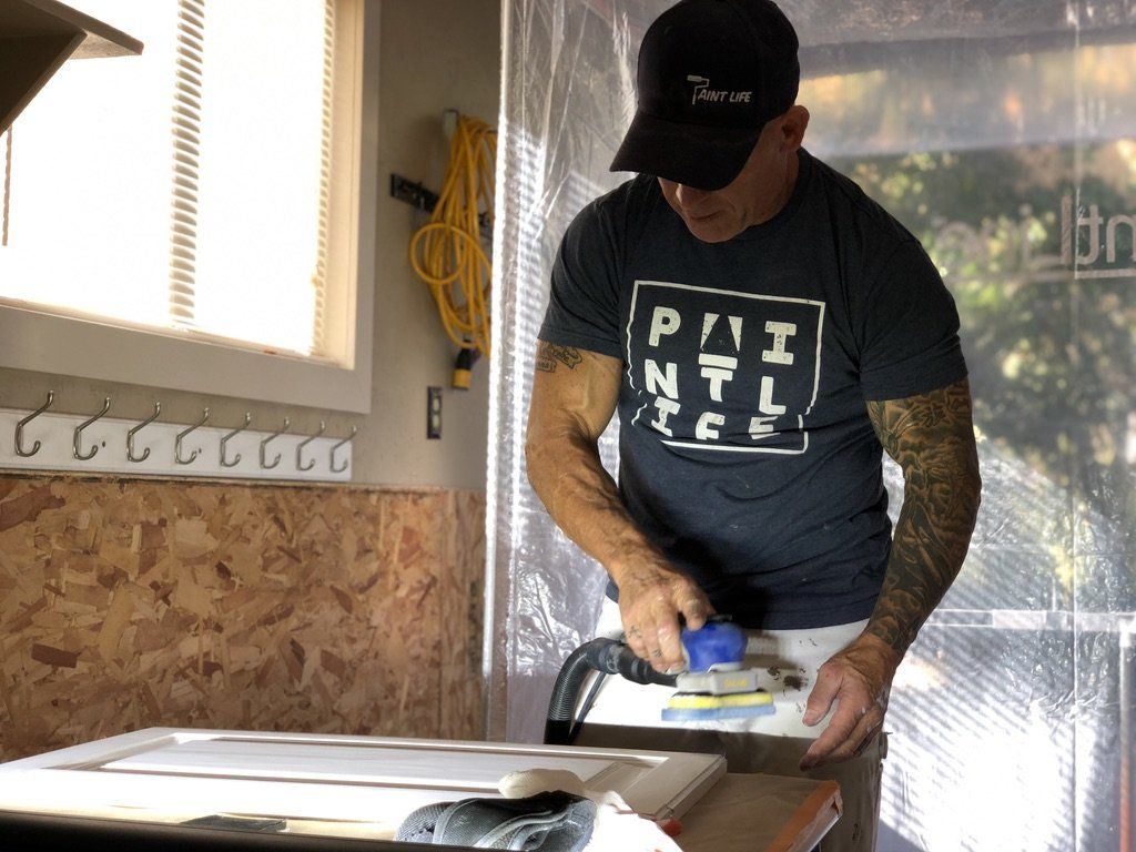 The Idaho Painter wearing a Paint Life tee is sanding a piece of wood.