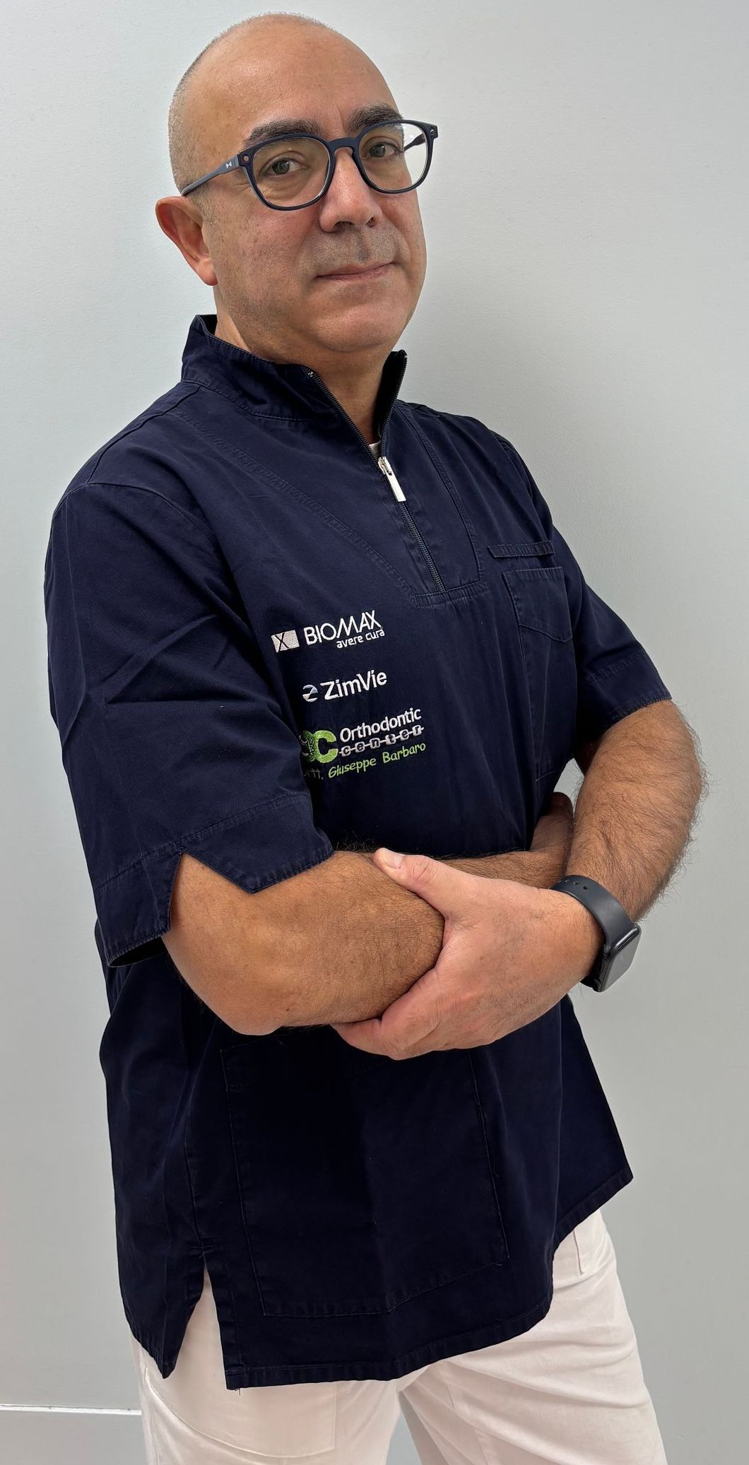 Un uomo calvo che indossa occhiali e una camicia blu è in piedi con le braccia incrociate.