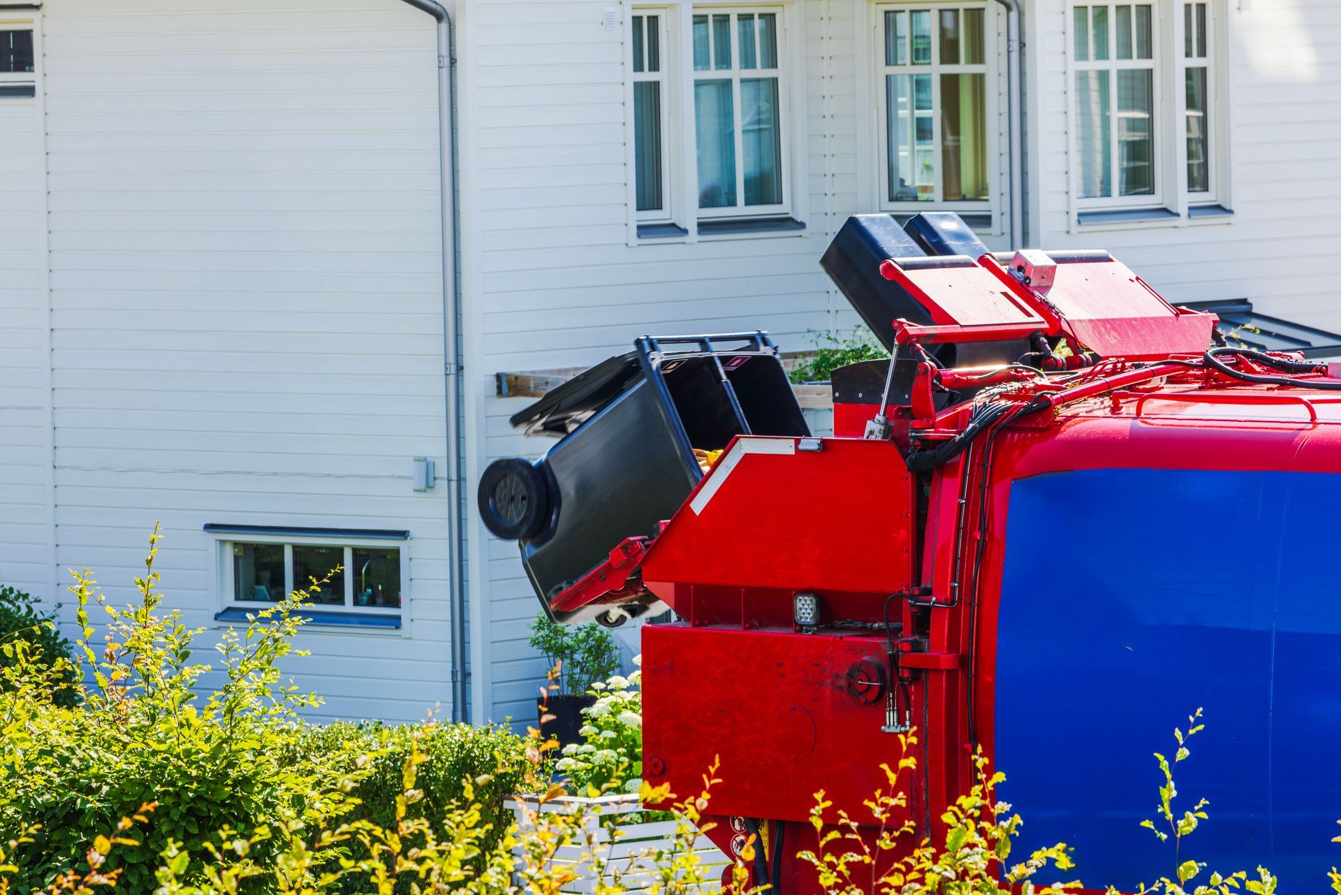 Red garbage truck lifting a black trash bin in front of a white house.