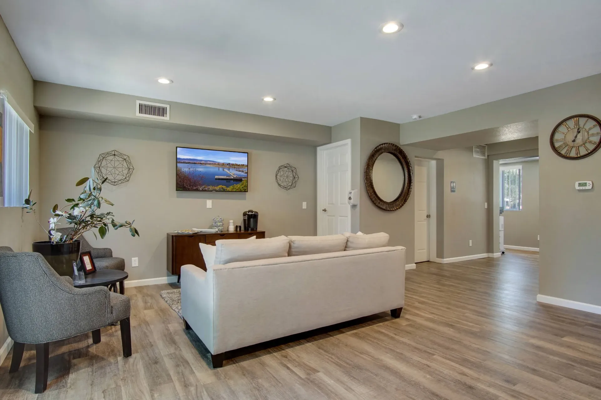 Living room in an apartment with a beige sofa, accent chairs, wall-mounted TV, and wood-look flooring.