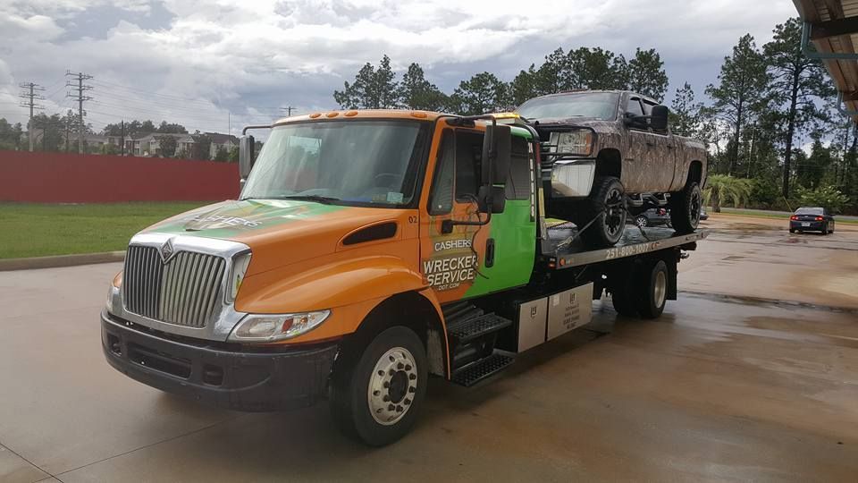 Orange tow truck carrying a damaged black pickup on a wet road near a red wall and trees