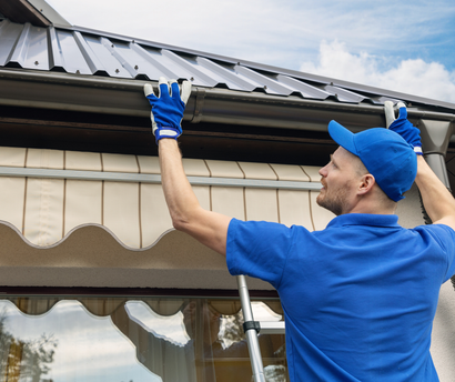 Man on ladder cleaning gutters, wearing blue shirt and gloves.