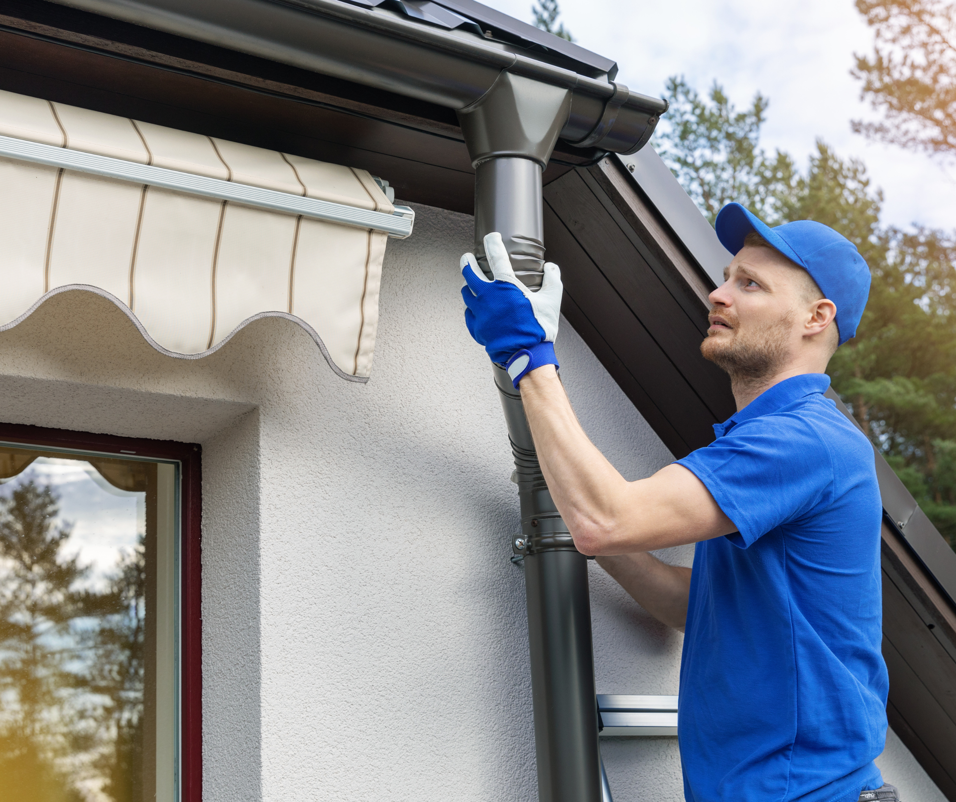 Person in blue uniform fixing a downspout on a house.
