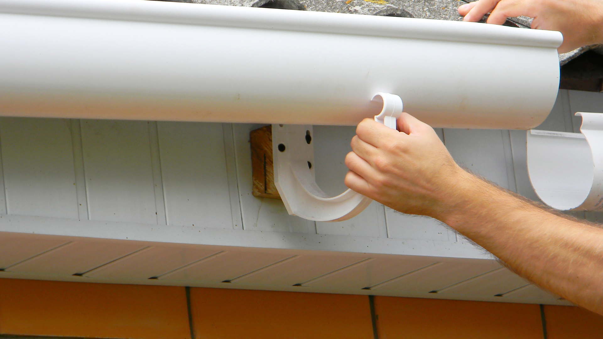 A person on a ladder adjusts a white plastic gutter section along the edge of a brick building.