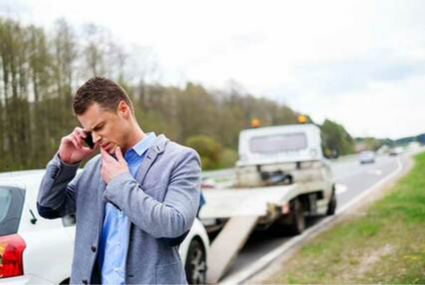 Picture of Guy at side of road on the phone while a tow truck is setting up to tow his car