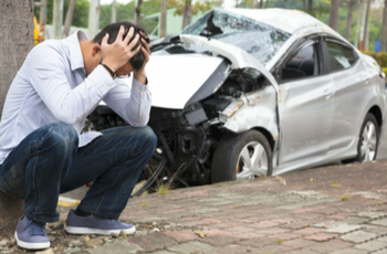 Picture of Guy with glasses hunched by tree with his white car very smashed up in background