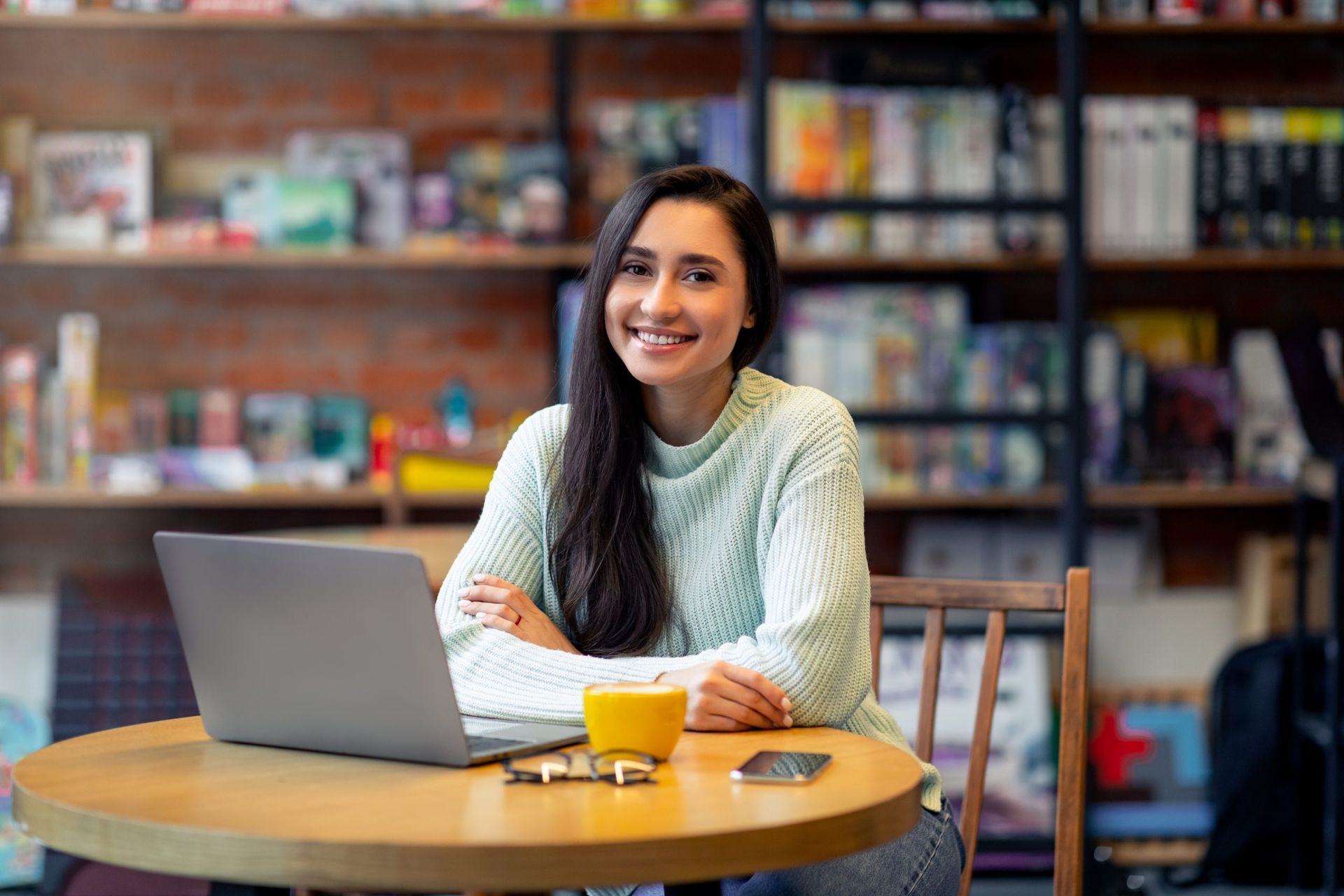 woman in green sweater smiling in a cafe. Laptop on table with glasses, phone and coffee cup. Board games and books in the background on shelves