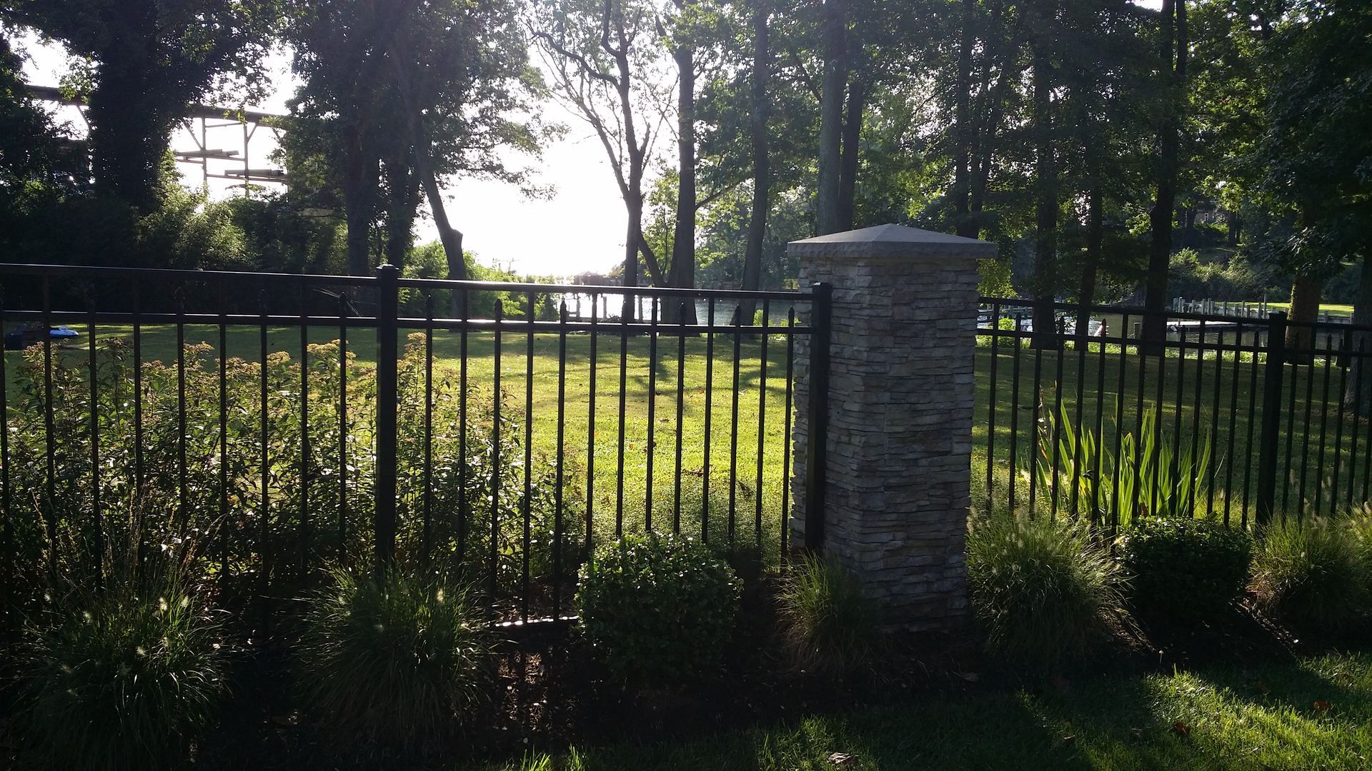Black metal fence with stone column in front of a grassy area with trees, bright sunlight.