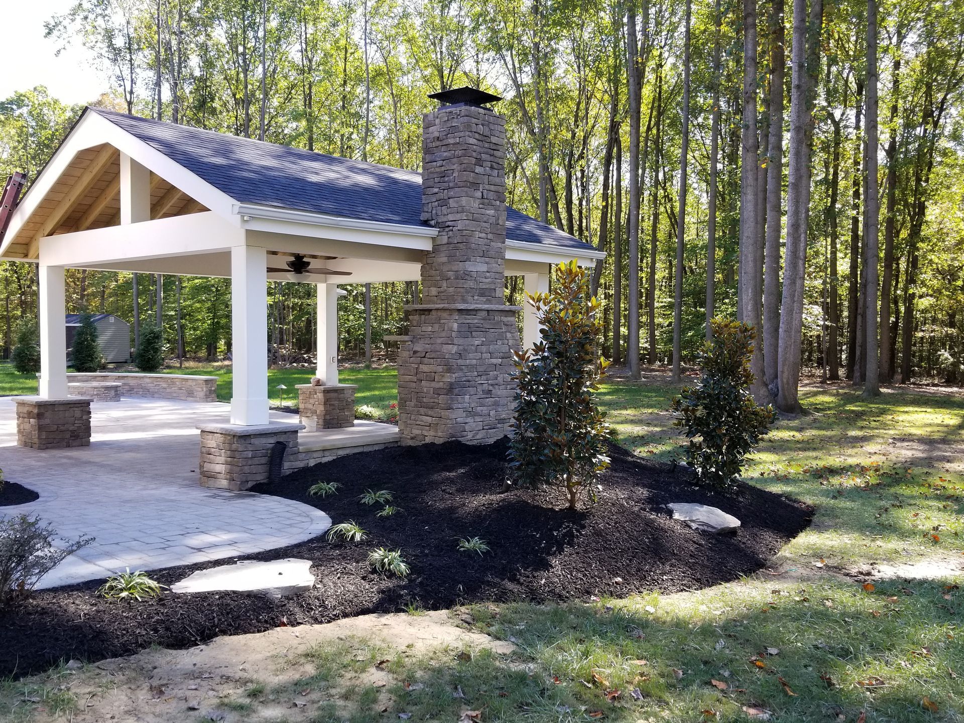 Pavilion with stone chimney and patio, surrounded by trees and dark mulch.