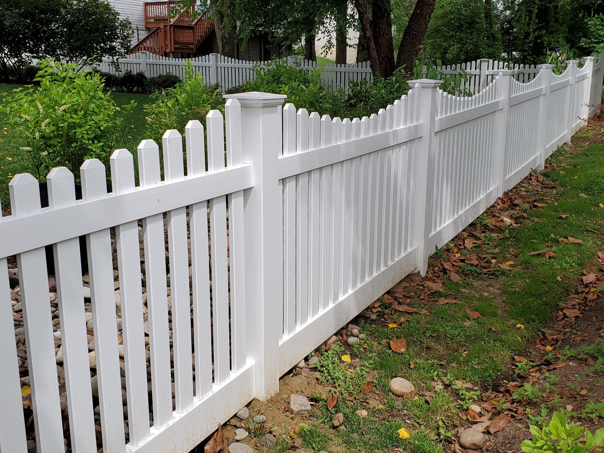 White picket fence in a grassy yard, curving in front of bushes and trees.