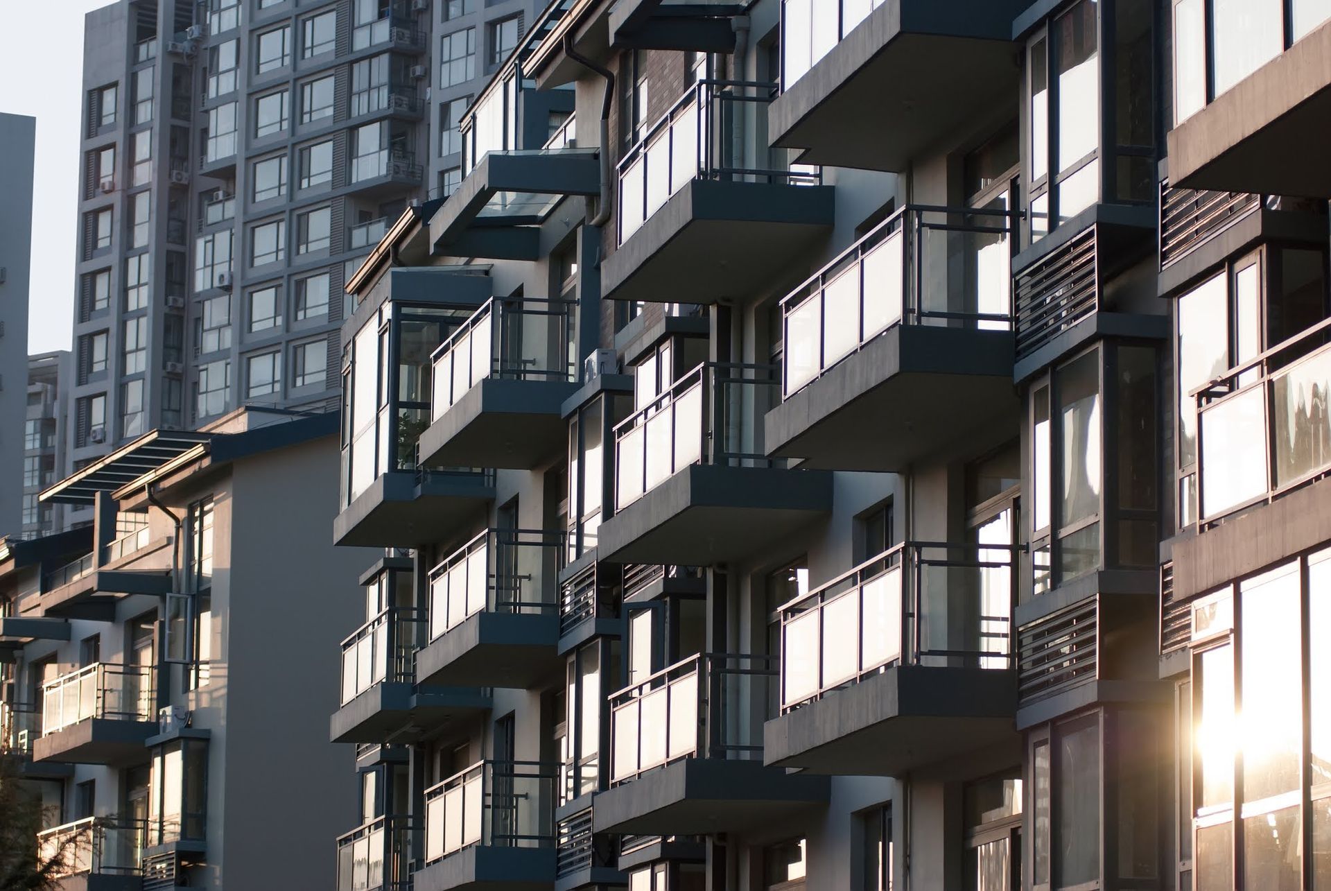 Row of modern apartment buildings with balconies, bathed in sunlight. Tall buildings in the background.