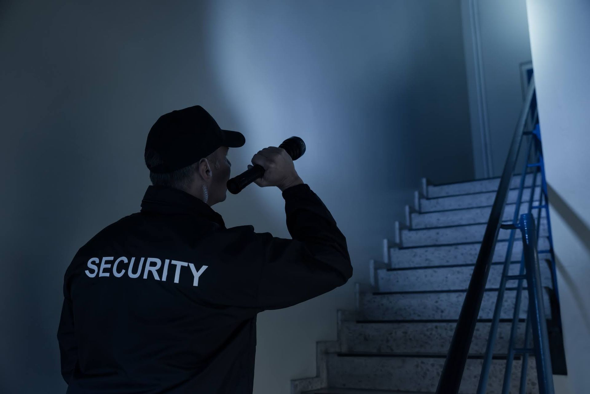 Security guard with flashlight in a dimly lit stairwell, wearing a black jacket labeled 