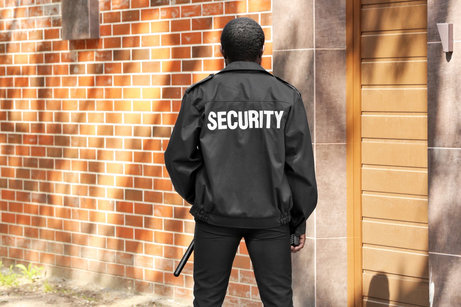 Security guard in black jacket standing on a city sidewalk near brick wall.