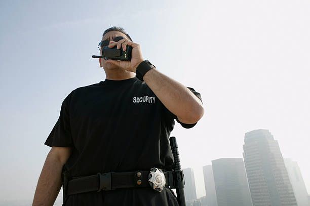 Security guard in black uniform using walkie-talkie with city buildings behind.