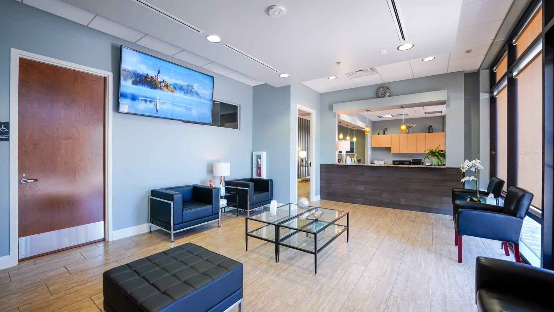 A waiting room with chairs , a coffee table and a flat screen tv.