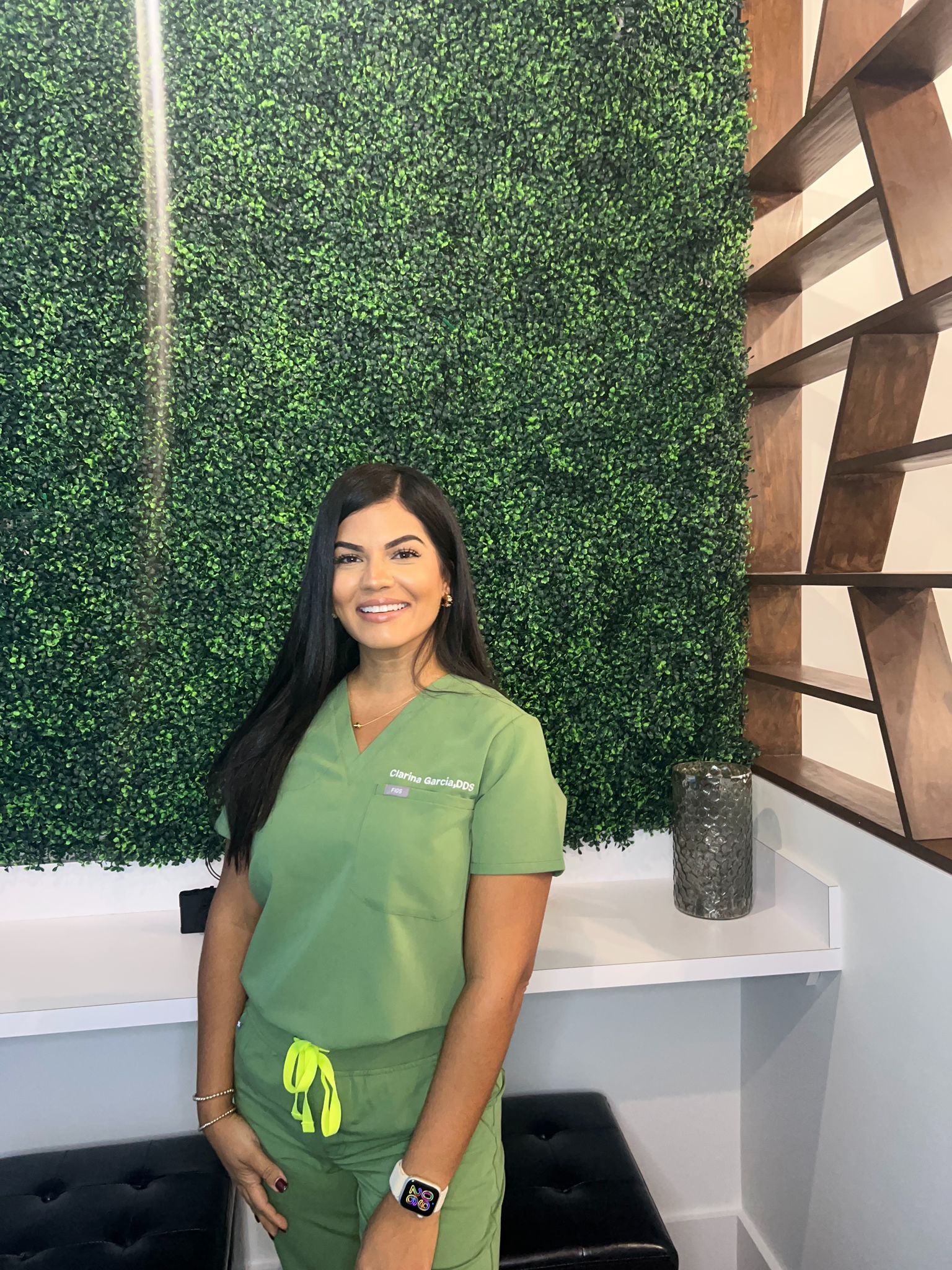 A female dentist wearing a white coat and a black scrub is smiling for the camera.