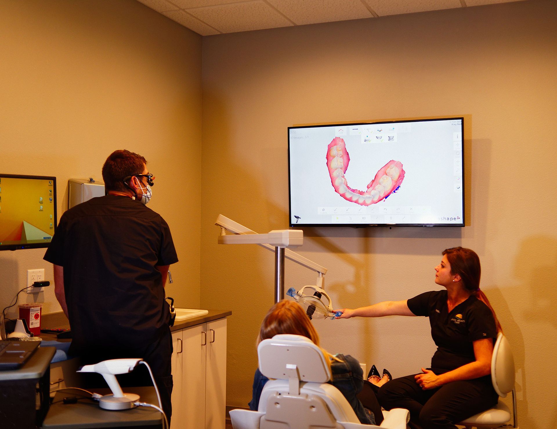 A woman is sitting in a dental chair while a man stands behind her.