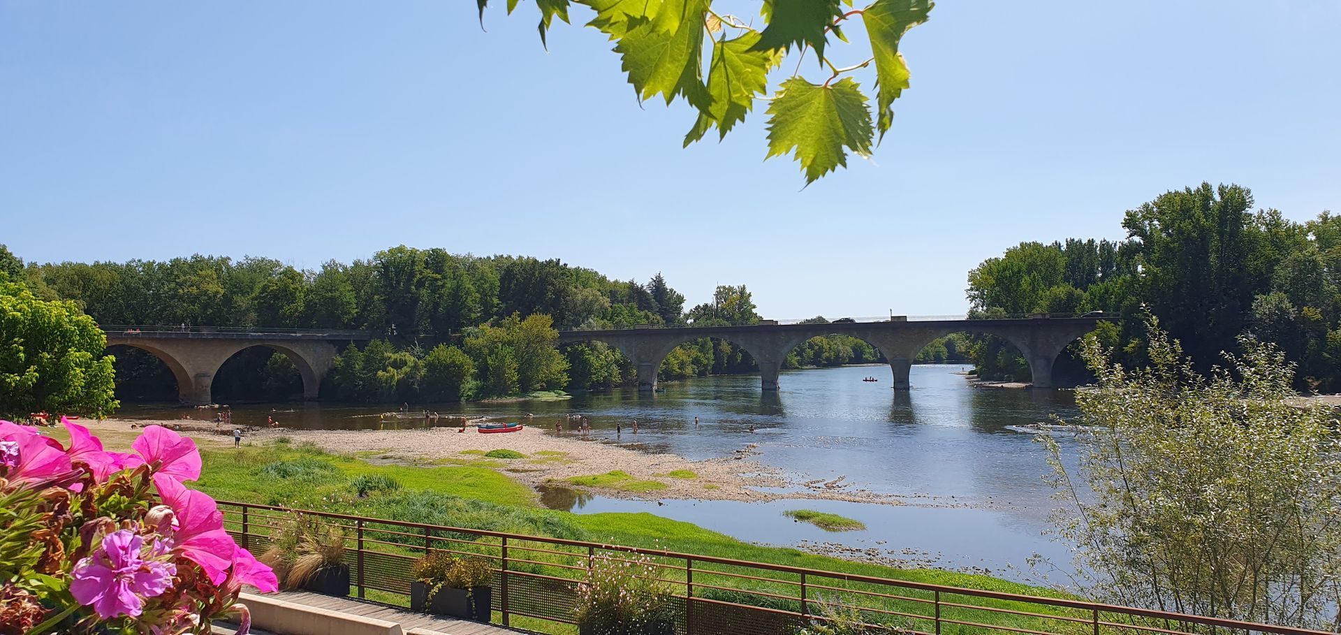Een stenen brug over een rivier op een zonnige dag, omlijst door groene bomen en roze bloemen.