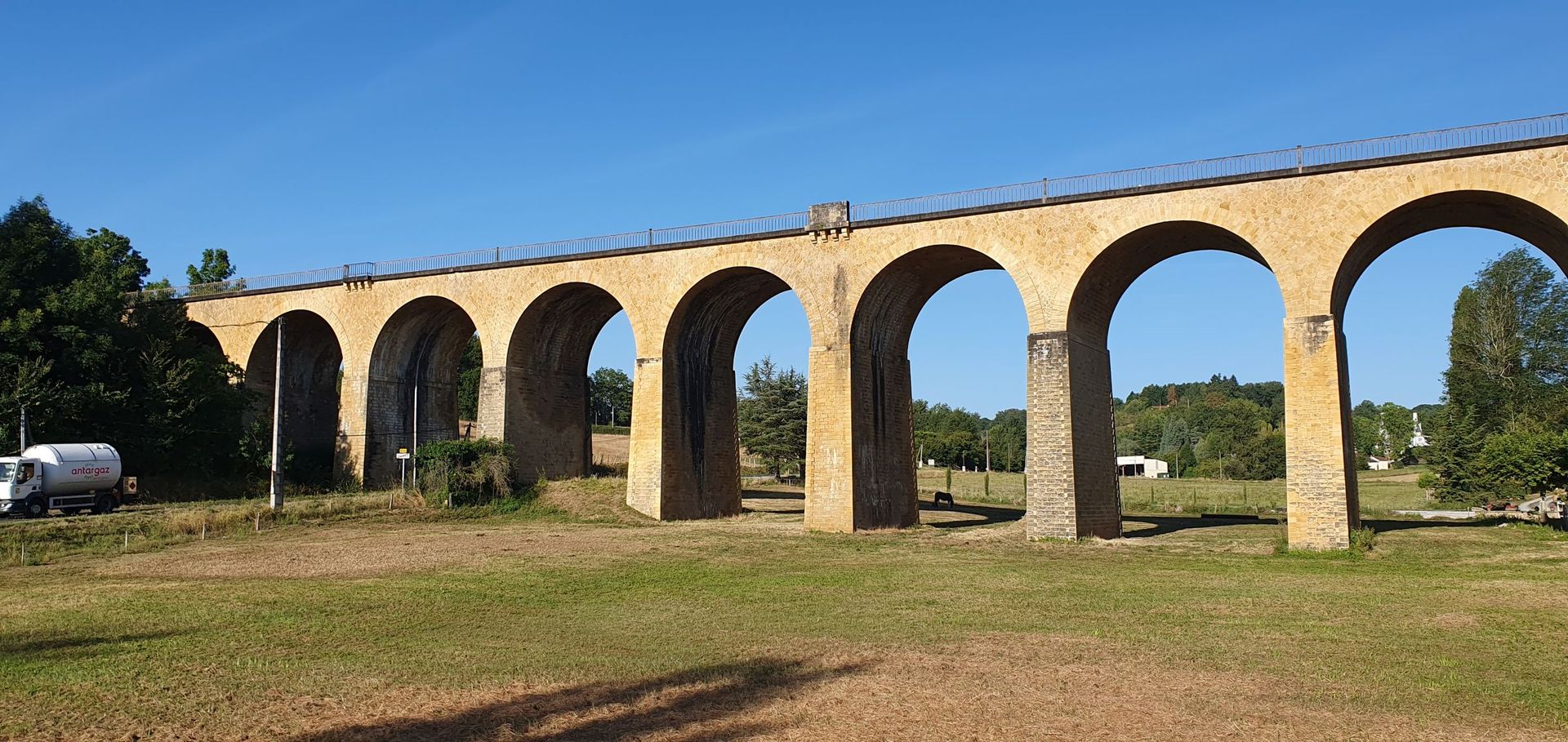 Een oud stenen viaduct voor treinen  overspant een grasveld onder een helderblauwe lucht.