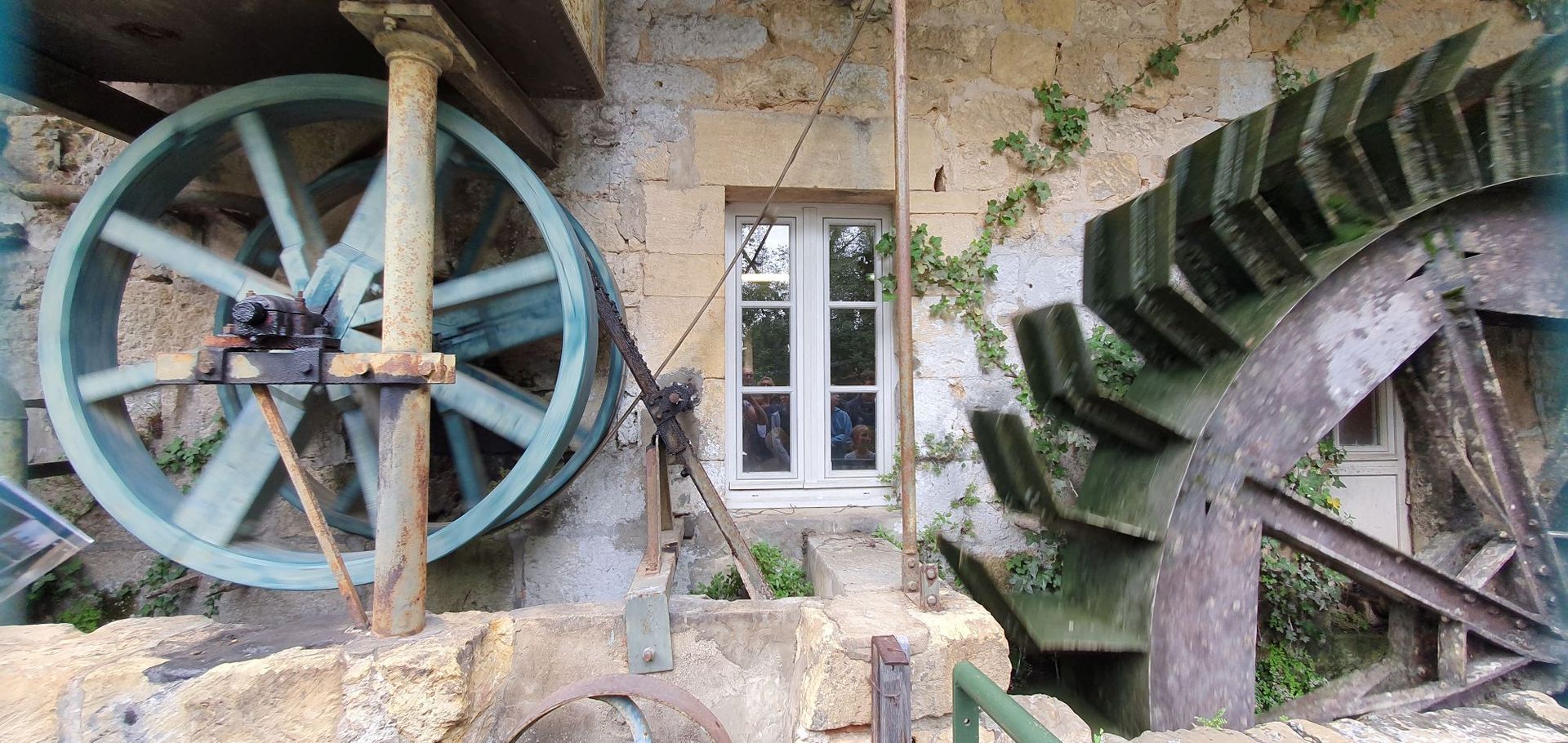 Een watermolen met twee grote wielen en een klein raam, in een stenen gebouw met groene begroeiing.