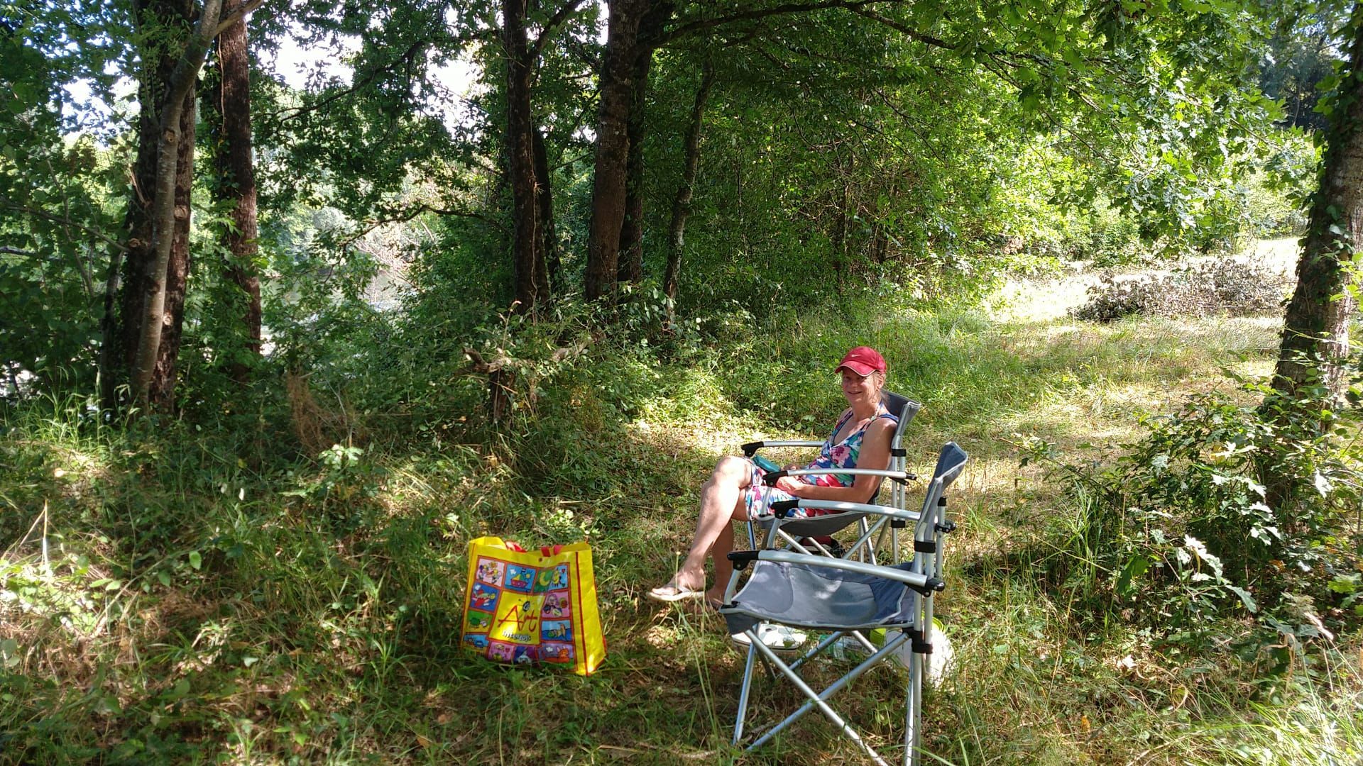 Woman sits in a folding chair in a shady outdoor area, red hat, colorful bag nearby.