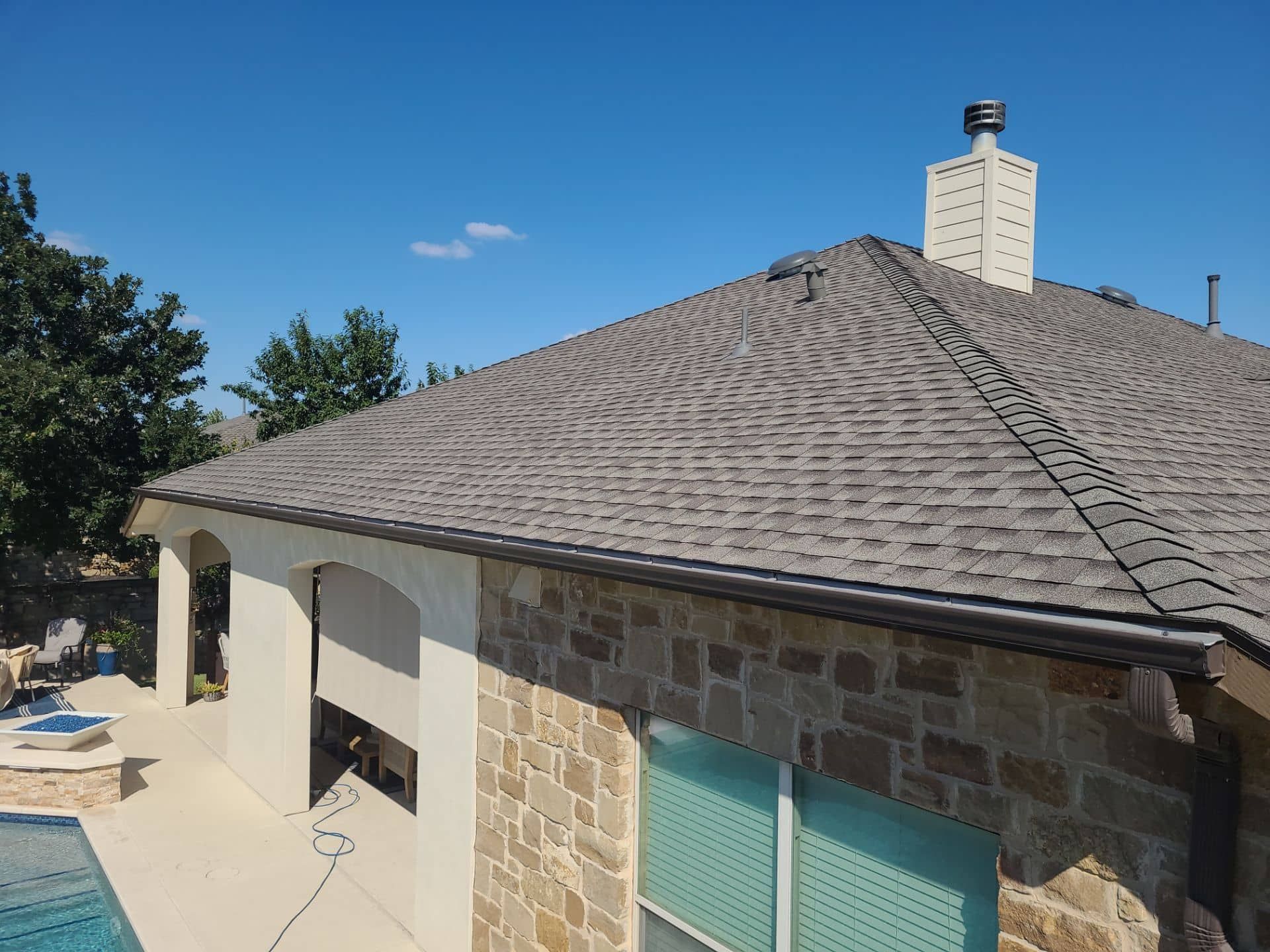 House roof with brown shingles, chimney, and stone and stucco walls, with a pool and blue sky visible.